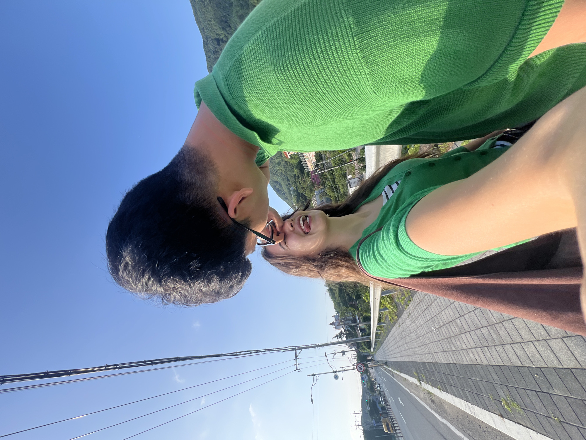 This is a vertical, selfie-style photograph featuring two young people, appearing to be a couple, in a close embrace. The man, on the left, has short, dark hair and is wearing a bright green sweater. The woman, on the right, has long, light brown hair and is wearing a green top with black and white stripes visible underneath. She is wearing glasses and her mouth is open in a happy expression, suggesting laughter or joy. They are positioned so their faces are very close, with the man leaning in towards the woman.
The background shows a clear, bright blue sky, indicating a sunny day. Below them, a landscape unfolds with green hills dotted with buildings, suggesting an elevated viewpoint overlooking a town or village. A paved pathway with distinct grey paving stones runs alongside a road, where a few cars are visible. Power lines stretch across the sky, supported by poles. A circular speed limit sign with "40" is visible on a pole, along with streetlights. The overall scene suggests an outdoor, possibly scenic location. The time of day appears to be daytime, given the bright sunlight and clear sky. The mood conveyed by the couple's interaction is affectionate and joyful. The image is taken from a high angle, looking down at the road.