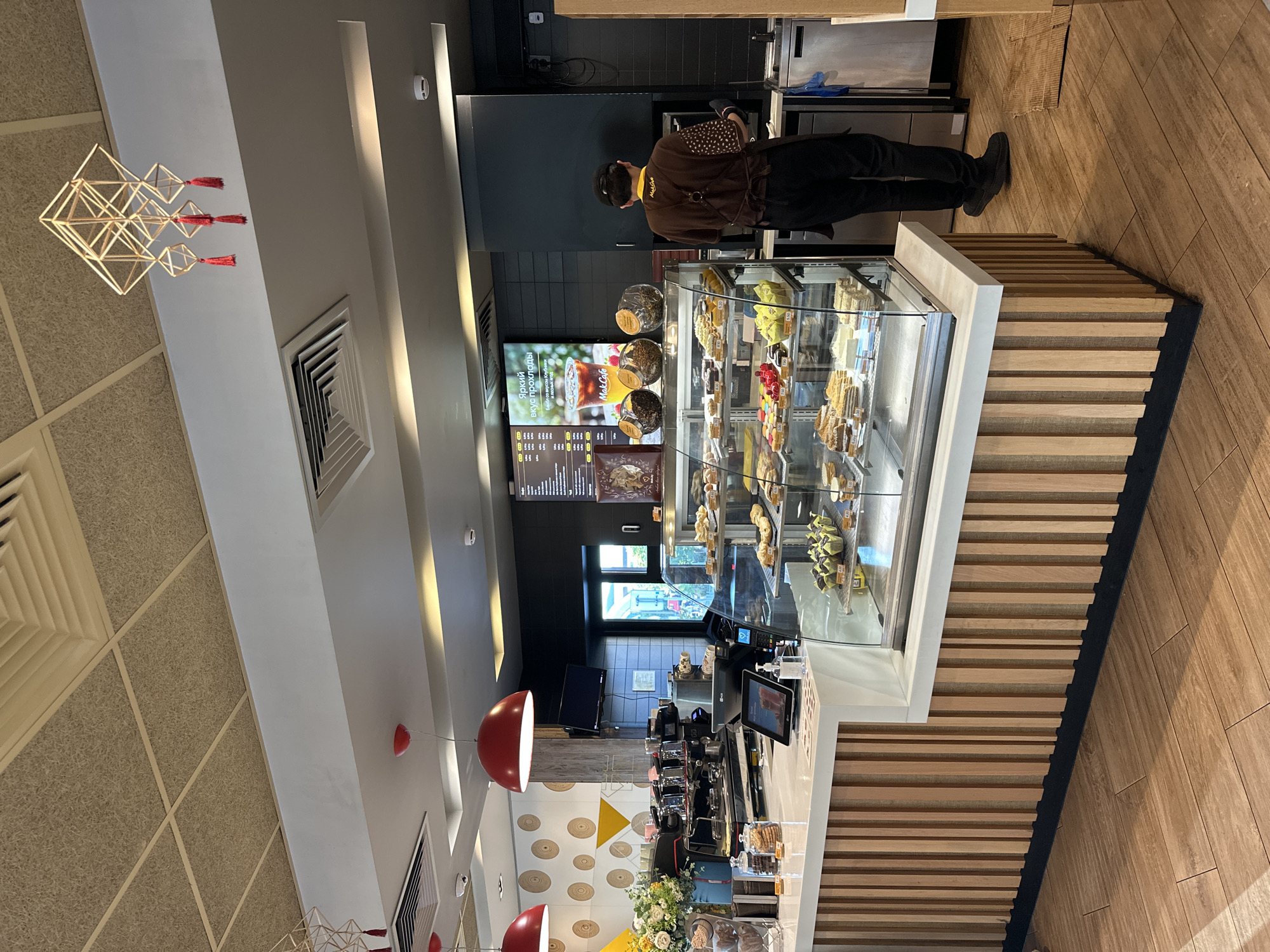 The image depicts the interior of a McDonald's McCafe located in Minsk, Belarus. The scene is a brightly lit, modern cafe with a wooden floor and light-colored walls. A counter displays a variety of pastries and baked goods in a refrigerated glass case. Behind the counter, a McDonald's employee wearing a brown uniform and an apron is visible. Coffee machines and other equipment are present on the counter. The cafe is decorated with geometric light fixtures and circular wall art. A digital menu board displays food and drink options. The overall atmosphere is clean and inviting, consistent with a fast-food restaurant environment.