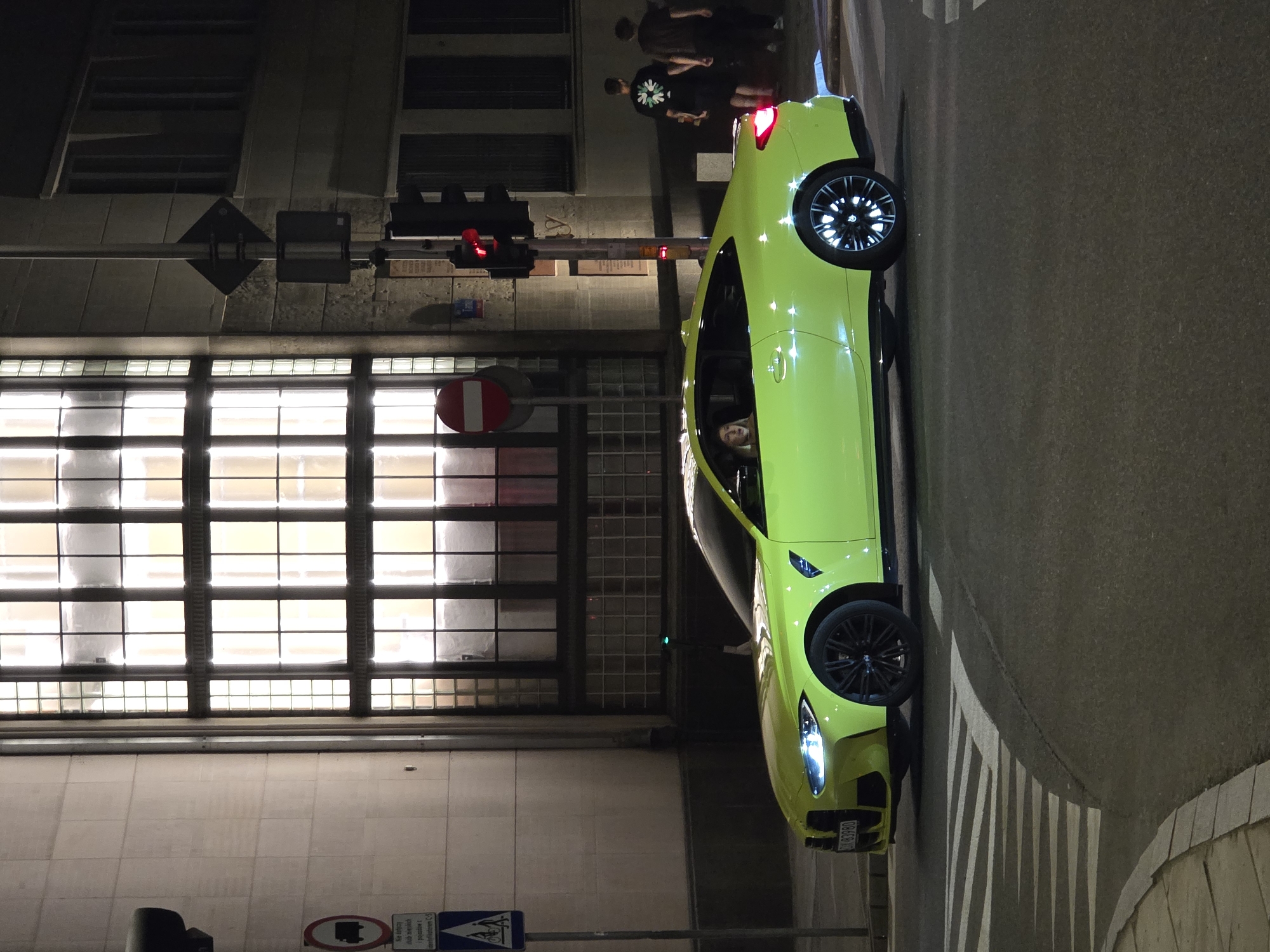 The image captures a neon yellow BMW M4 parked at a street corner in Warsaw, Poland, at night. The car is positioned on the right side of the frame, facing left. A person with blonde hair is visible in the driver's seat, looking forward. The car's headlights and distinctive wheels with illuminated elements are illuminated.
Behind the car, a building with large, multi-paned windows emits a warm, yellow light. A traffic light shows a red signal, and adjacent to it are traffic signs, including a circular sign with a red border and a horizontal white bar, indicating "No Entry" for vehicles, and a blue square sign with pictograms of people on bicycles and walking. Two individuals are walking away from the car on the sidewalk in the background. The street surface is asphalt, with white painted pedestrian crossing lines visible in the foreground, indicating a crosswalk. The scene is lit by streetlights and the interior lights of the building, creating a nighttime urban atmosphere.