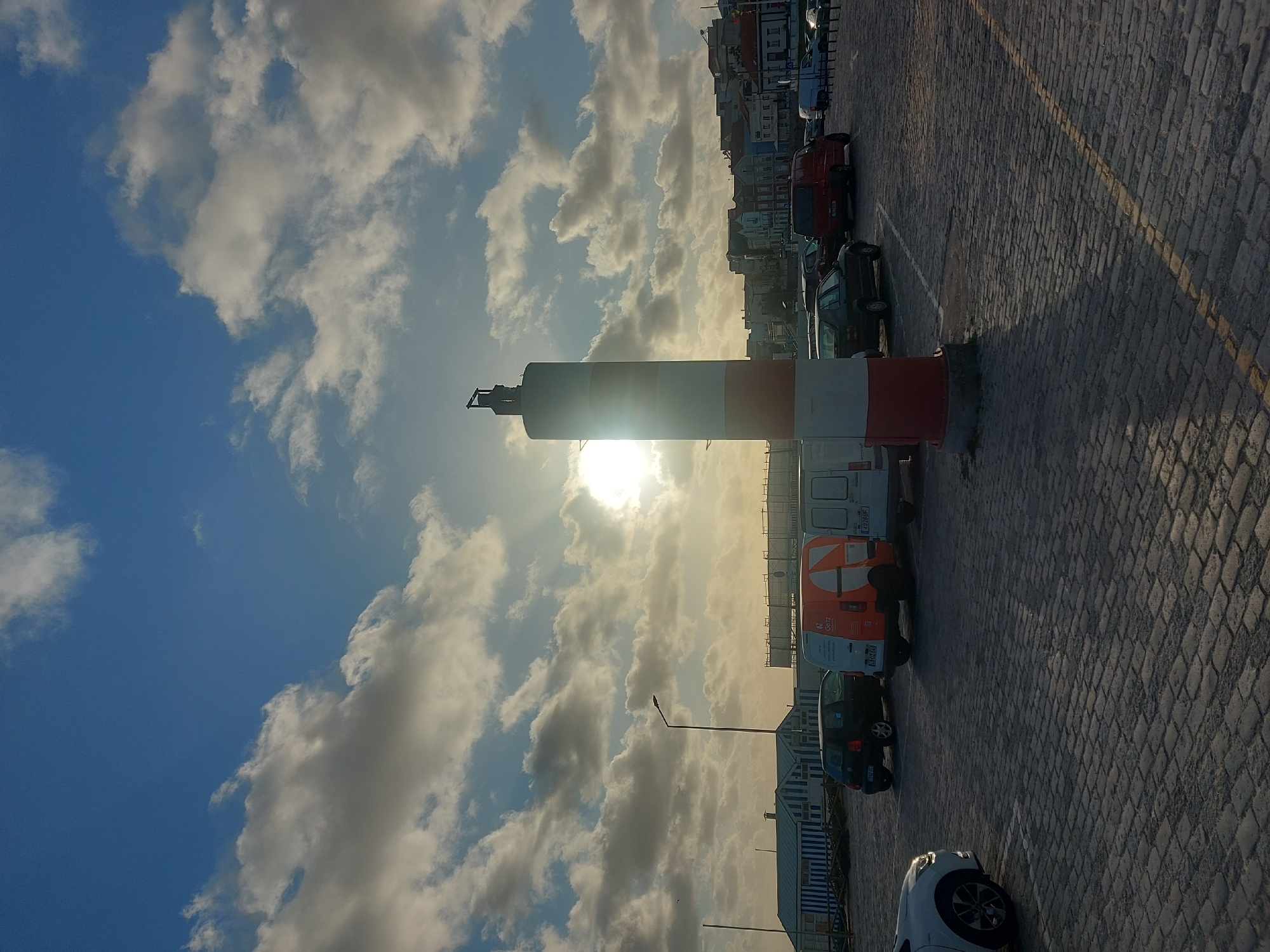 The image presents a view of the Paredão da Praia da Claridade promenade in Figueira da Foz, Portugal. A cylindrical navigational beacon painted in alternating white and red bands is positioned prominently in the foreground, partially obscuring the sun. The sky is filled with a mixture of blue patches and billowing white clouds. Along the promenade, parked vehicles include vans and cars, partially lining the edge of the cobblestone pavement. Beyond the vehicles, a row of buildings is visible. The paving stones of the promenade stretch out, marked by a single yellow painted line.