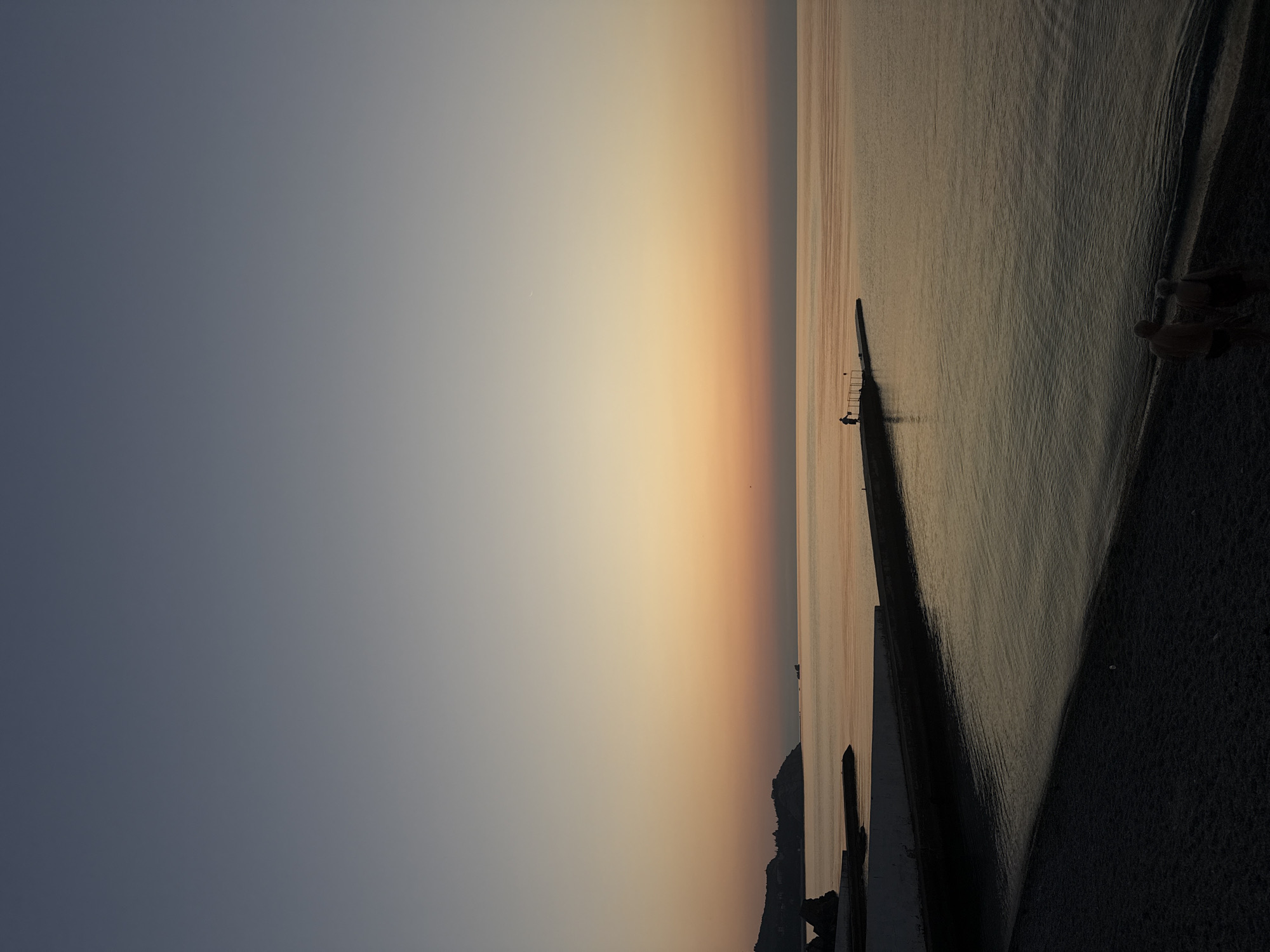This is a vertical, low-angle, full shot of a beach and the sea at dusk in Partenit, Ukraine. The sky transitions from a pale blue at the top to a soft orange near the horizon. The sea is calm and reflects the colors of the sky. In the distance, a man stands on a pier, and two figures, an older couple, walk along the pebble beach in the foreground. The beach is dark, and the water's edge is visible. A concrete embankment runs along the left side of the image, separating the beach from the sea. Hills with buildings are visible in the far distance to the left.