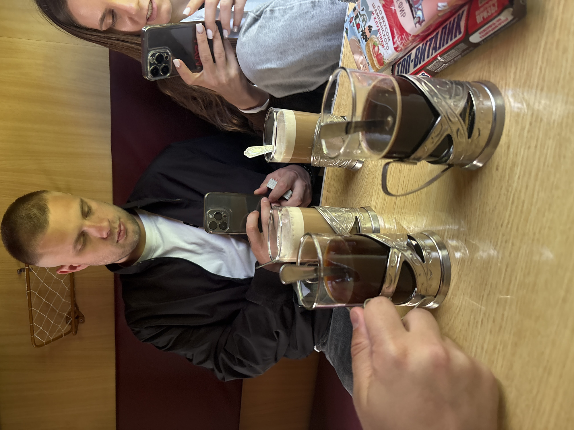 In a train cabin, two individuals are seated at a wooden table. One person is holding a smartphone, while the other is stirring a drink in a glass with a metal holder. A second glass with a metal holder sits beside it. A packaged food item lies on the table. The seat is upholstered in a dark red material, with a rack on the wall. Based on the location, this is likely a scene from a train journey through Тарноўскі сельскі Савет, Belarus.