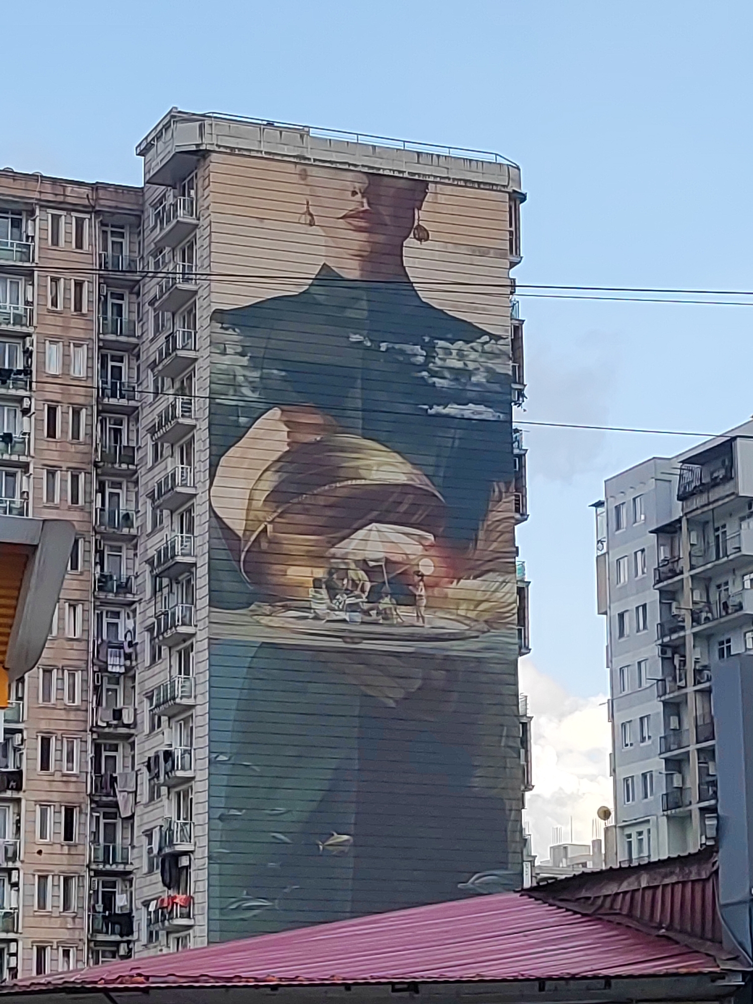 The image shows a mural painted on the side of a tall, multi-story building in Batumi, Georgia. The mural depicts a woman's upper torso and head, facing slightly to the left. She is wearing a dark outfit and large earrings. Her head is adorned with a patterned headscarf. Below her chest, the mural transitions to an underwater scene with several dolphins and fish swimming in a greenish-blue expanse. In the mid-section of the mural, there appears to be a smaller scene with figures under a beach umbrella, suggesting a beach or coastal setting. The building itself is light brown and has a grid of windows and balconies. To the left of this building is another residential building with a similar architectural style. To the right, another, more modern, light-colored building is partially visible. A section of a red-tiled roof is in the foreground at the bottom of the image. The sky is blue with scattered clouds. The mural is a prominent feature of the urban landscape.