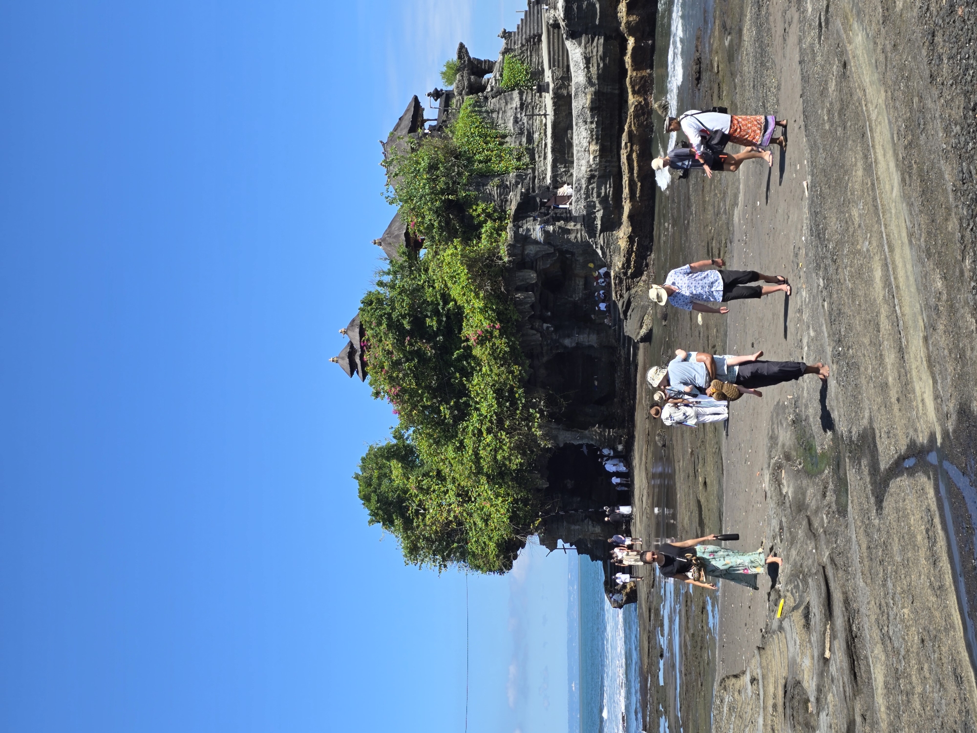 A bright, sunlit coastal landscape is captured in this image, featuring a prominent temple complex built upon a rocky offshore outcrop. The image is composed with a vast, clear blue sky dominating the upper portion.
The central subject is a large, dark grey rock formation, situated to the left of the frame, adorned with lush green foliage and trees. Perched atop this natural structure are several traditional temple buildings, characterized by their multi-tiered, dark pagoda-style roofs. Stone steps and pathways are visible winding up the rock face, suggesting access to the temple.
In the foreground and extending across the middle ground is a dark, wet sandy or rocky shore, indicating a low tide that exposes a wide expanse of the seabed. The ocean stretches out to the right and behind the rock formation, with gentle white-capped waves breaking near the shore.
Several people are scattered across the wet ground. In the immediate foreground, a man in a light blue patterned shirt, black shorts, and a straw hat walks to the left. Behind him, another individual wears a white top, a patterned sarong, and a head covering. Further back, a woman in a dark top and light patterned pants stands. Many other people, some dressed in white traditional Balinese attire and sarongs, are gathered closer to the base of the rock formation, possibly exploring an alcove or cave opening.
The scene takes place on a bright, clear day with abundant sunlight and a cloudless blue sky, suggesting fair weather conditions during midday or early afternoon. The architecture and setting strongly imply the location is Tanah Lot Temple in Bali, Indonesia. No visible text appears in the image.