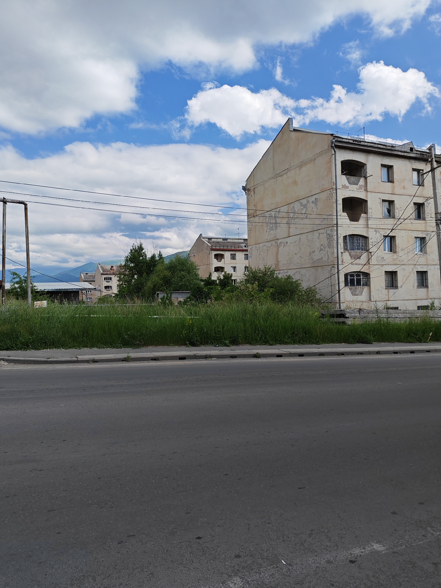 The image depicts a street view in Vanadzor, Armenia, on a cloudy day. In the foreground, a gray asphalt road stretches across the bottom of the frame. A concrete sidewalk borders the road, separating it from a patch of tall, green grass. Behind the grass, several apartment buildings are visible. The closest building on the right is a light-colored, multi-story structure with a weathered facade, showing signs of peeling paint. It has several windows and balconies, some of which are enclosed. Another similar building is partially visible to its left, and further in the distance, more apartment buildings are seen nestled among trees. Utility poles and power lines crisscross the scene. In the background, mountains with a hazy blue appearance are faintly visible against the sky, which is dotted with white clouds. The overall impression is of a residential area in a city, with elements of urban infrastructure and natural surroundings.