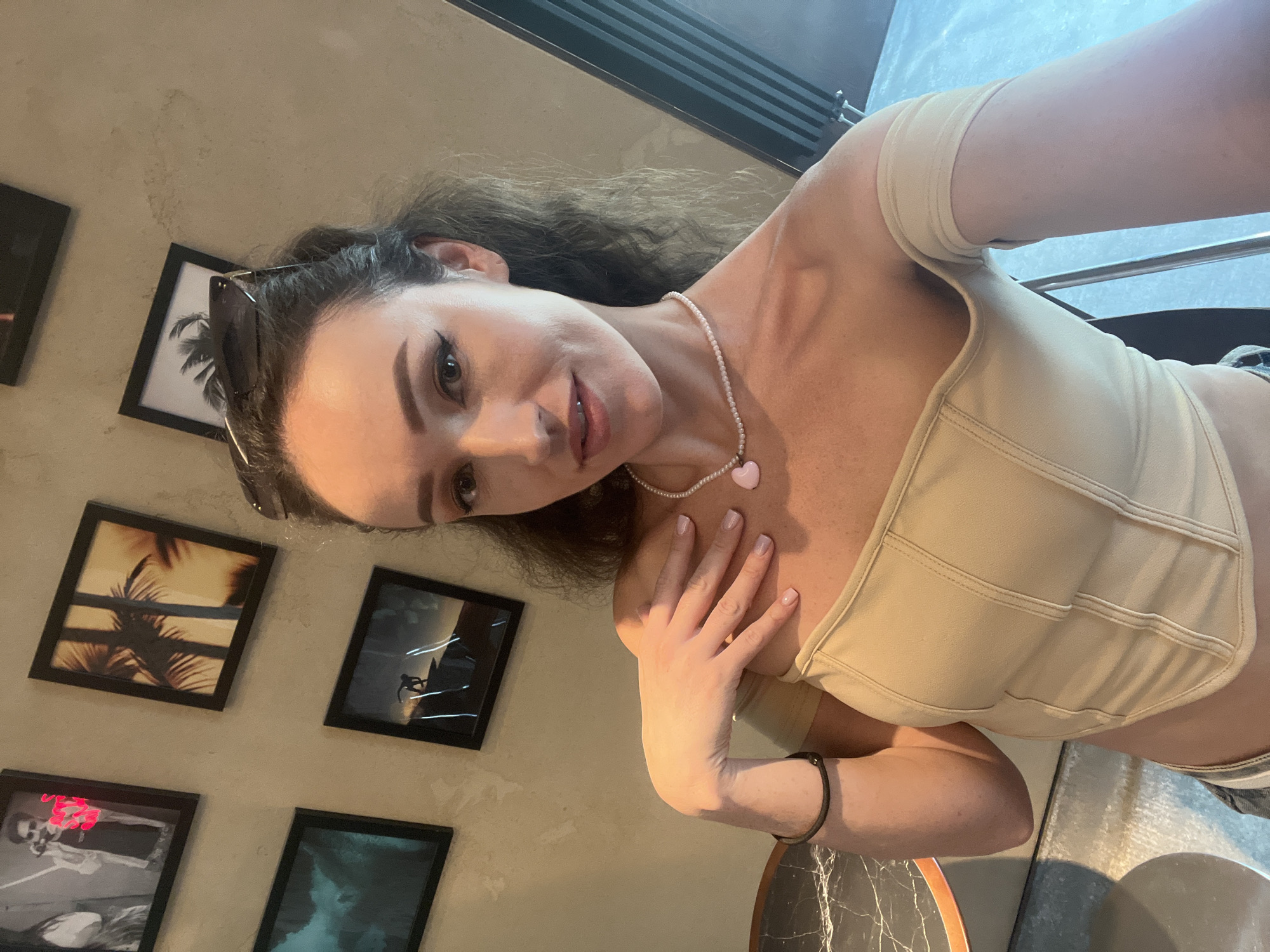 A young woman is taking a selfie in a room with framed pictures on the wall. She is wearing a beige off-the-shoulder top and a pearl necklace with a pink heart pendant. Her sunglasses are perched on her head. The room appears to be indoors, possibly a cafe or a restaurant, with a marble-topped table visible in the foreground. The background shows several framed photographs of various scenes, including what appears to be a performer on stage, palm trees, and a surfer. There is no indication that this location is in Kazan, Russia.