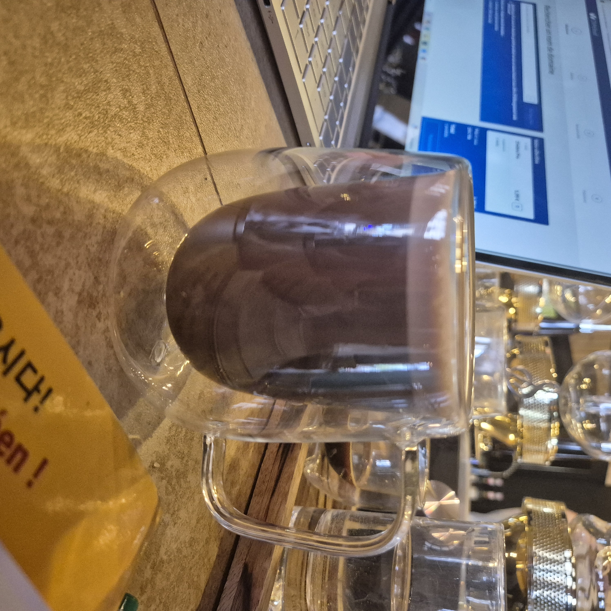 The image shows a glass mug filled with a dark liquid, likely coffee or chocolate, sitting on a table near a laptop. A yellow object with Korean writing is visible on the left. In the background, there are other glasses and some wood. The scene appears to be indoors, possibly in a cafe or workspace. While I cannot definitively confirm the location in Strasbourg, France, based on the visual information, the presence of Korean writing suggests a location where there may be a Korean cultural influence, which could include a cafe or business in Strasbourg. The presence of a laptop implies someone is working or browsing the internet.