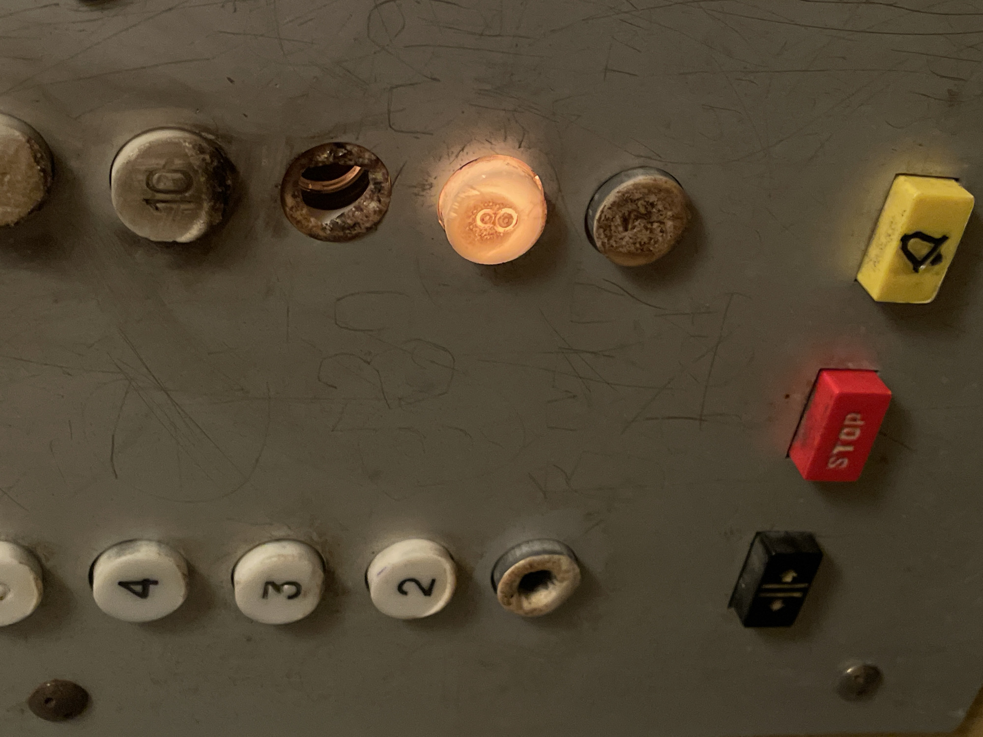 The image shows a close-up view of an elevator control panel. The panel is made of metal and has several buttons, some of which are illuminated. From top to bottom on the left side, there are buttons labeled with numbers 4, 3, and 2. Below these are two unlabelled, worn buttons. On the right side, there are buttons labeled with numbers 10 and 8. The button labeled 8 is illuminated with a warm, orange light. Below the button labeled 10, there is an empty, rusted circular opening. Further down on the panel are three smaller buttons: a black button with two yellow vertical lines and a diamond shape, a red "STOP" button, and a yellow button with a bell symbol. The metal panel is scratched and appears worn. The setting is likely an older building's elevator. There is no information in the image to suggest a location in Tbilisi, Georgia.
