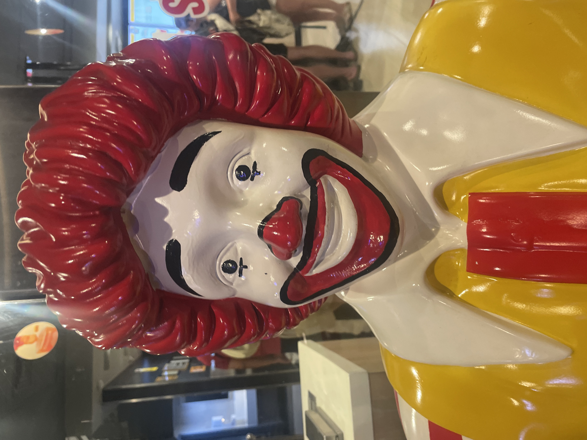 The image shows a statue of Ronald McDonald with his iconic red hair, white face, and clown smile. The statue is positioned inside, possibly in a restaurant or establishment related to McDonald's. Behind Ronald McDonald, there is a blurred view of an interior setting with a window or glass surface, reflecting light. The background includes hints of objects and surfaces, giving the impression of a commercial space. The overall lighting seems bright and artificial.
The location is not evident from the image, but the prompt asks to include Denpasar, Indonesia. Thus, the statue is located inside an establishment in Denpasar, Indonesia.