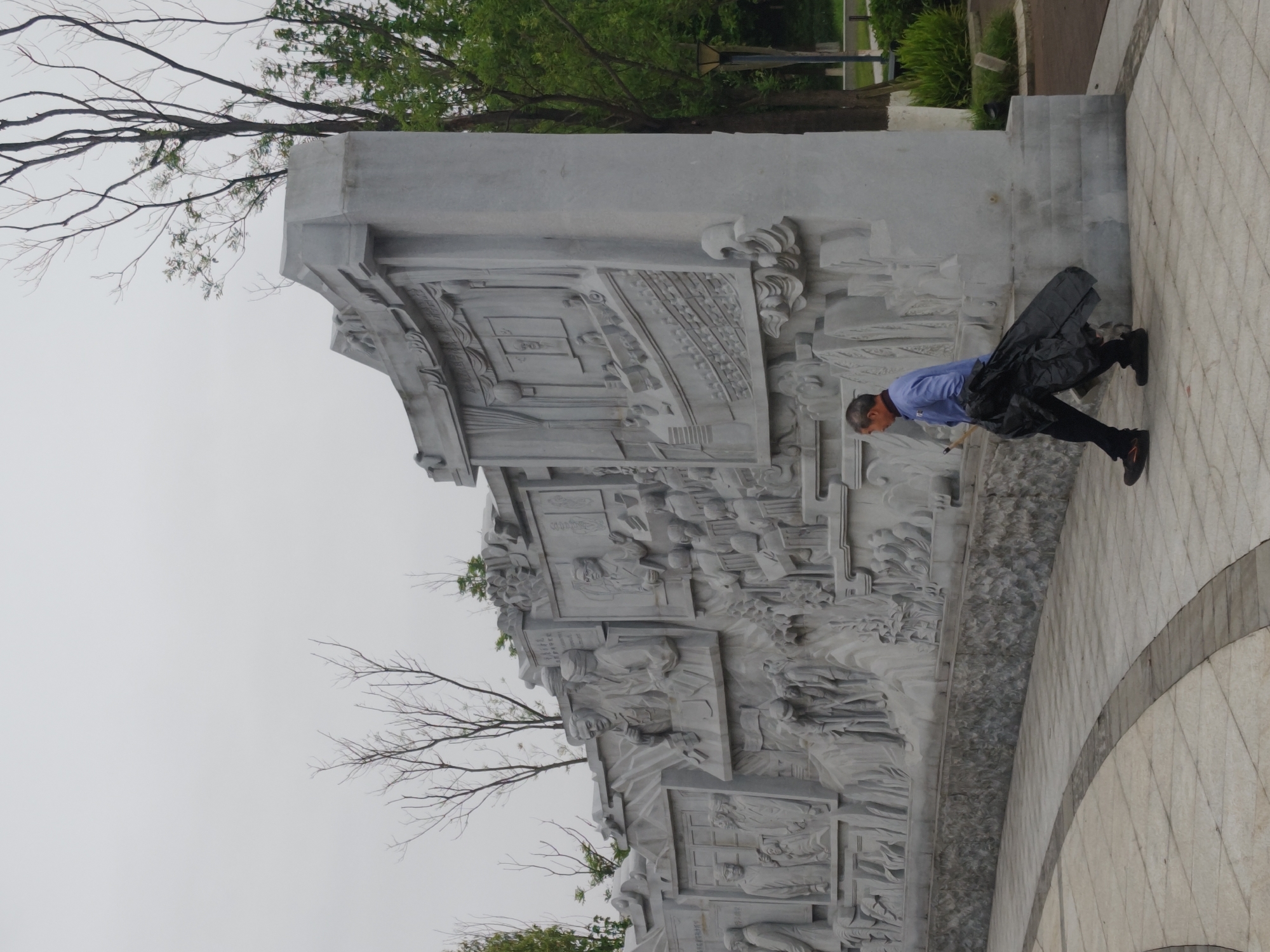 The image, taken in Chongqing High-tech Zone, China, displays a large, elaborate grey stone relief sculpture and an adult male engaged in an activity. The multi-paneled sculpture dominates the left side of the frame, depicting numerous figures and detailed scenes, including architectural elements such as buildings and interior settings like classrooms. Faint Chinese characters are visible on plaques and banners within the carved scenes, although they are largely unreadable due to distance and resolution.
In the foreground and to the right, an older adult male with short, graying hair is captured in a dynamic pose. He wears a blue long-sleeved shirt, dark trousers, and dark shoes, one of which has a distinct red sole detail. He is bent forward at the waist with his knees flexed, holding a long, thin object that resembles a stick or a broom. His posture suggests he is either performing a sweeping motion on the paved ground or practicing a form of martial arts or exercise, such as Tai Chi with a staff.
The scene is set outdoors, likely in a public park or plaza, indicated by the patterned paved ground in the foreground, featuring lighter and darker grey tiles arranged in a curved design. The sky is overcast, providing soft, diffused lighting. Bare tree branches and green foliage are visible in the background and extending above the sculpture, suggesting a natural environment surrounding the monument. Distant trees, bushes, and a streetlamp are faintly discernible beyond the sculpture on the right.