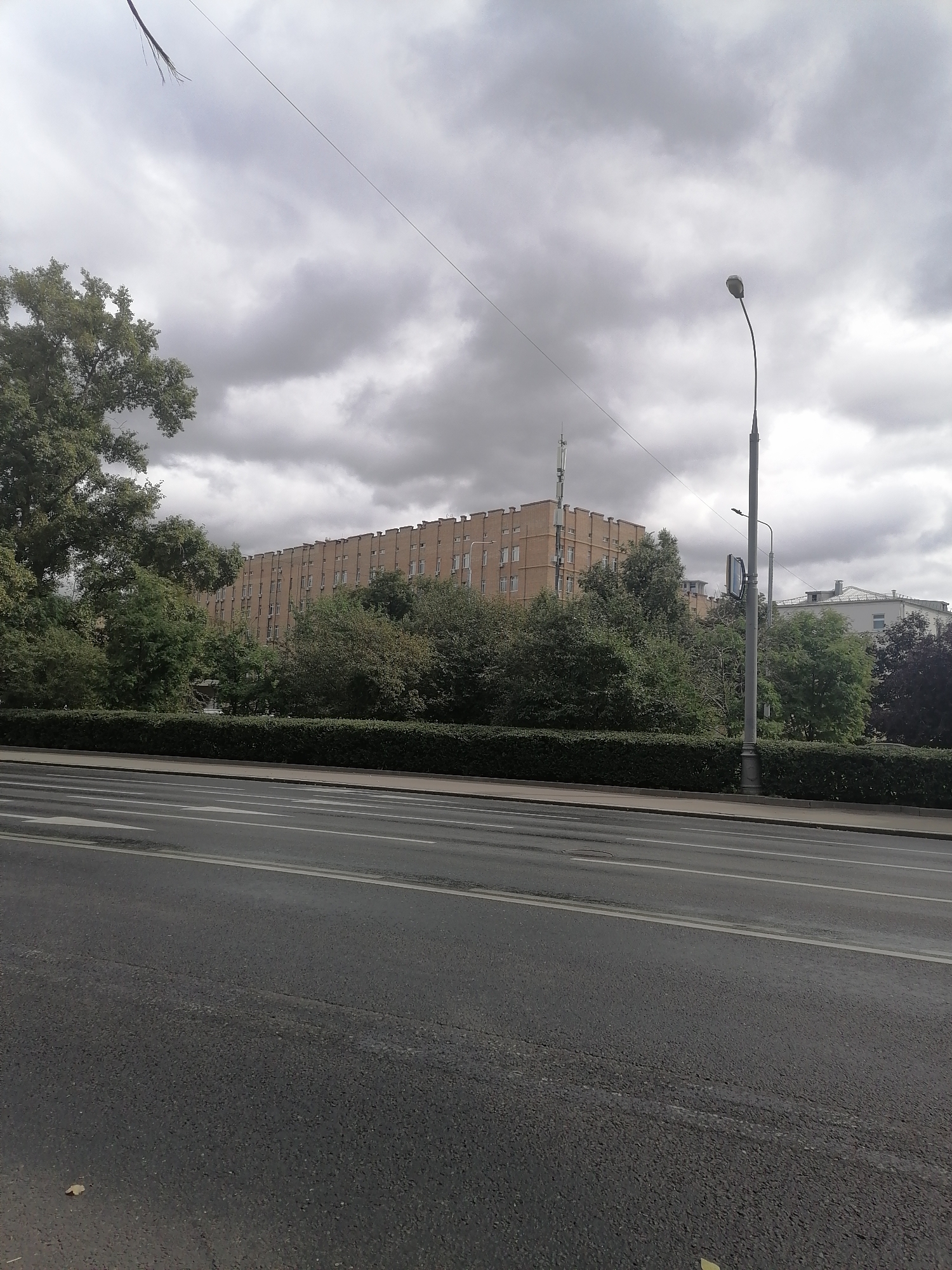 A wide shot captures a street scene under a heavily clouded sky. In the foreground, a grey asphalt road features white lane markings. Beyond the road is a neatly trimmed green hedge. Behind the hedge, a long, beige brick building with numerous windows stretches across the frame. Trees with green foliage are interspersed in front of and around the building. A tall, grey lamppost stands to the right of the frame. In the background, other buildings and more trees are visible. The sky is filled with dense, dark grey clouds.