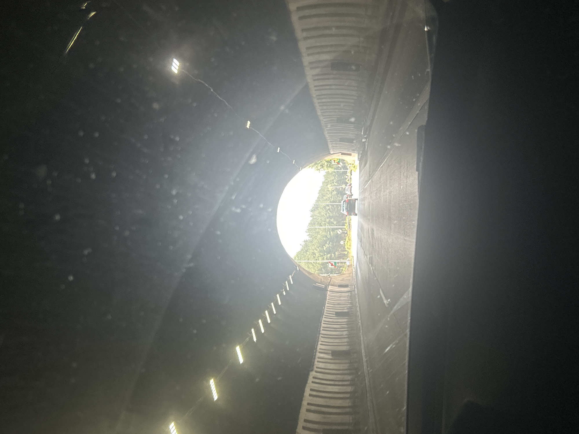 The image shows the interior of a tunnel, looking towards its exit. The tunnel is dark and lined with lights that are illuminating the path. On the right side of the tunnel, there is a concrete wall with a ridged texture. The left side of the tunnel also appears to be concrete, with lights embedded in it at intervals. The tunnel is wide and appears to accommodate vehicular traffic.
At the end of the tunnel, daylight is visible, with trees and some traffic on the road outside. There are several cars visible near the exit, suggesting that this is a road tunnel. The lighting from the exit creates a strong contrast with the darkness of the tunnel interior, with a bright, circular opening. There are some dust or water particles visible in the air within the tunnel, catching the light.
The scene depicts the experience of driving through a tunnel. The time of day appears to be daytime, given the bright light at the exit. There is no specific indication of weather conditions, other than the need for artificial lighting within the tunnel. The location is identified as Dolna Vasilitsa, Bulgaria. There is no visible text in the image. The overall mood is one of transition, moving from darkness to light.