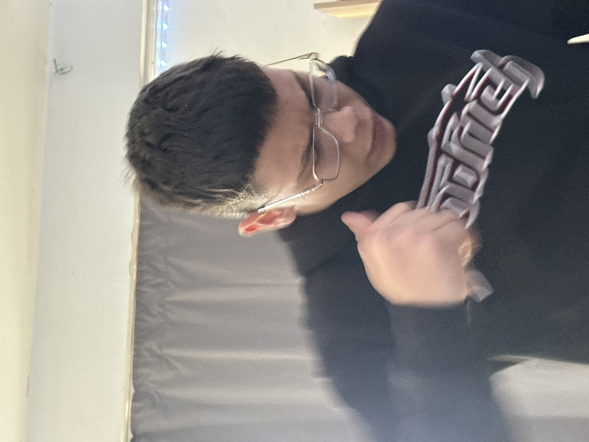 An indoor close-up shot captures a young person, likely male, from the chest up, looking intently downwards at an object they are holding. The person has short, dark hair and is wearing clear-framed rectangular glasses. They are dressed in a dark, possibly black, long-sleeved top or hoodie.
Their hands are visible near the center of the frame, holding a stylized gray or silver necklace or pendant. The pendant features intricate, ornate lettering that appears to spell out the word "SUPREME." The person's expression is focused, with their mouth slightly ajar, suggesting concentration on the item.
In the background, a light-colored wall is visible, with a section on the right featuring a string of small, bright LED lights. To the left, another light surface is present. Below the person's right arm, a crumpled or folded gray fabric, possibly a curtain or sheet, makes up the lower background. The overall lighting suggests an indoor setting, possibly an evening or night scene, given the presence of decorative lights. The location context provided is Troyan, Bulgaria.
