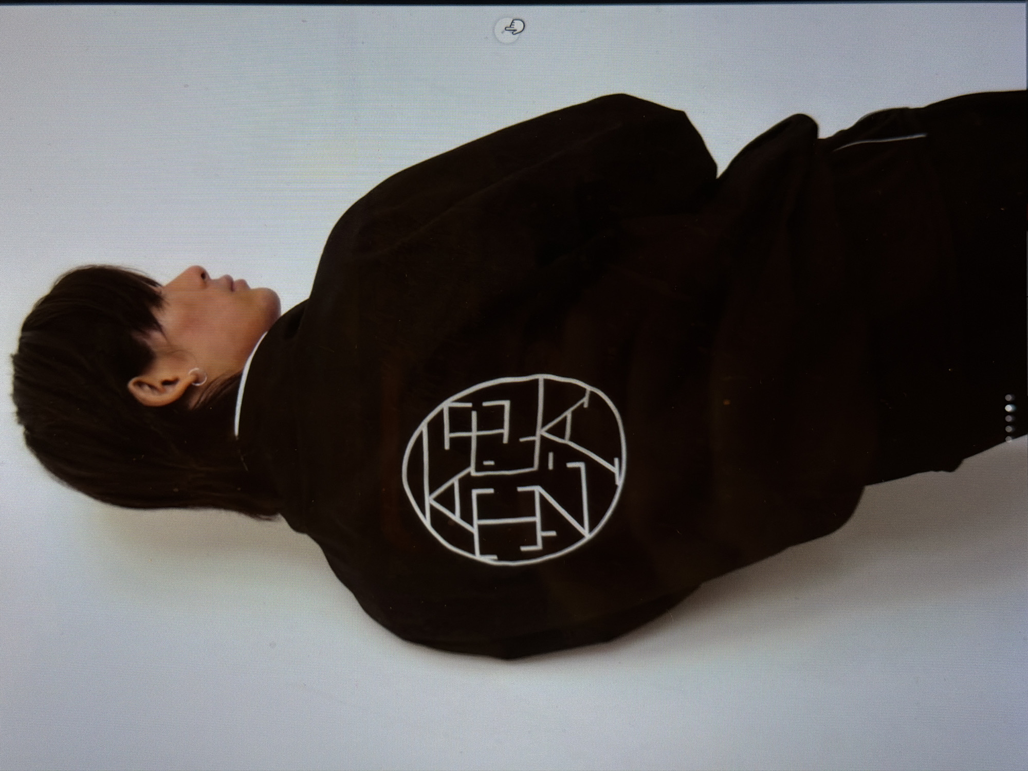 A person with short black hair, wearing a black jacket with a white circular design on the back, lies on a white surface. The design appears to contain the letters "KEN" within the circle. The person is lying on their side, facing away from the camera. The background is a plain white surface. It is not possible to determine the location.