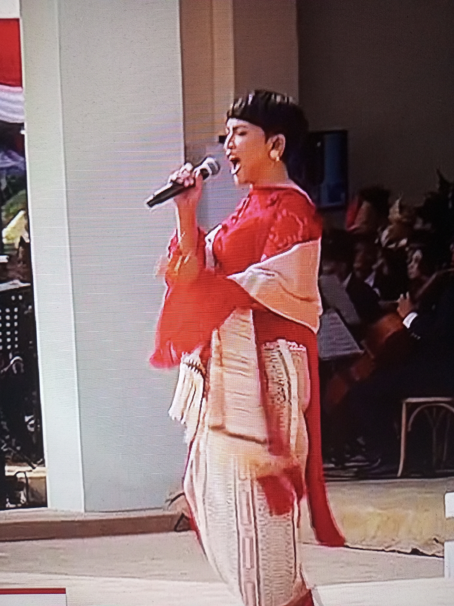 A woman with dark hair and a red top is singing into a microphone. She is wearing a light-colored sash and a patterned skirt. She is on a stage in front of a white pillar. In the background, there is an audience and a band. The image appears to be taken from a screen.
