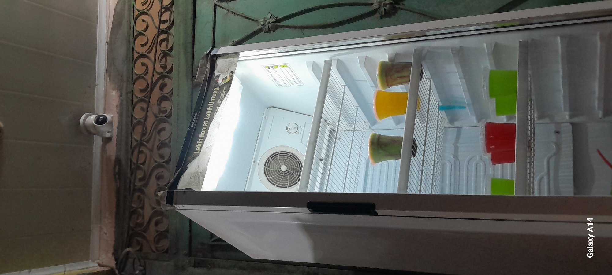 An open refrigerator is the primary subject of this indoor, eye-level, medium shot. The refrigerator door is on the left side of the frame, and the interior is illuminated. Inside, on the second shelf from the top, there are three cups of brightly colored drinks. The leftmost and rightmost cups contain a green beverage with darker green swirls, while the middle cup holds a yellow beverage. On the shelf below, there are two red cups and two green cups, all with clear plastic lids. A blue plastic stirrer is visible on the second shelf from the bottom. The refrigerator's internal fan is visible on the back wall of the top compartment. A security camera is mounted on the ceiling above the refrigerator. The background shows a wall with decorative metalwork and part of a green metal gate. The image is tagged with "Galaxy A14". The location is identified as Keudegeudong, Indonesia.