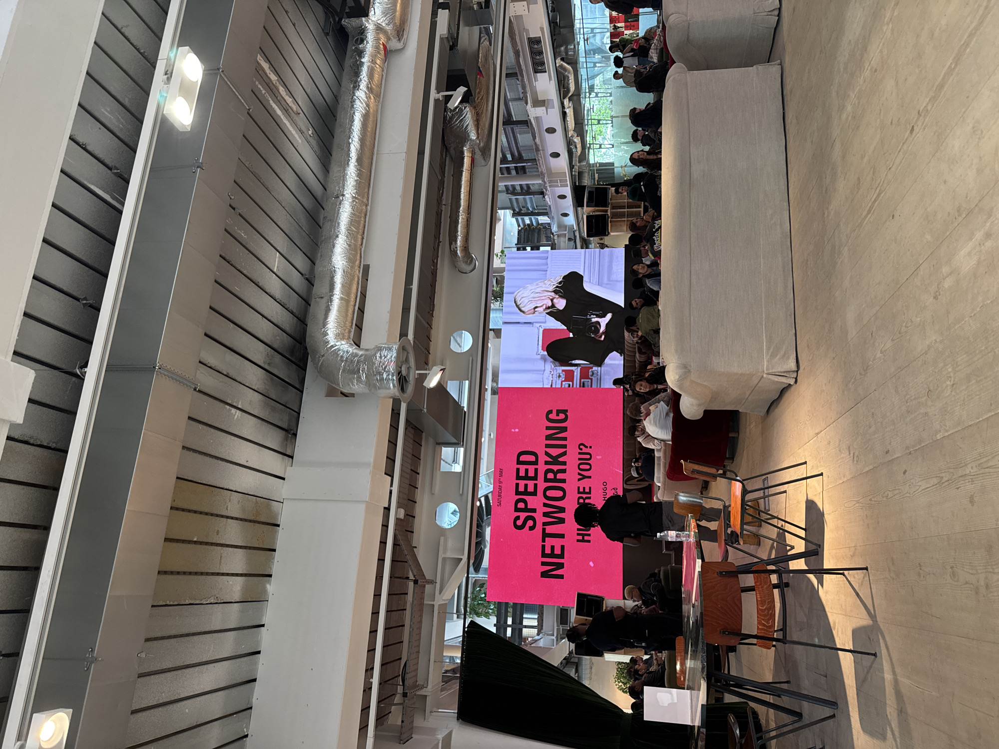 The image depicts an indoor event occurring in London, United Kingdom. Attendees are gathered, and a large pink banner with the text "SPEED NETWORKING" is displayed. A screen behind the banner shows an image of a person holding a camera. The setting appears to be a spacious, industrial-style venue with exposed ventilation systems and a high ceiling with overhead lighting. Furniture includes chairs and sofas for seating. People are standing and sitting, seemingly participating in the event.