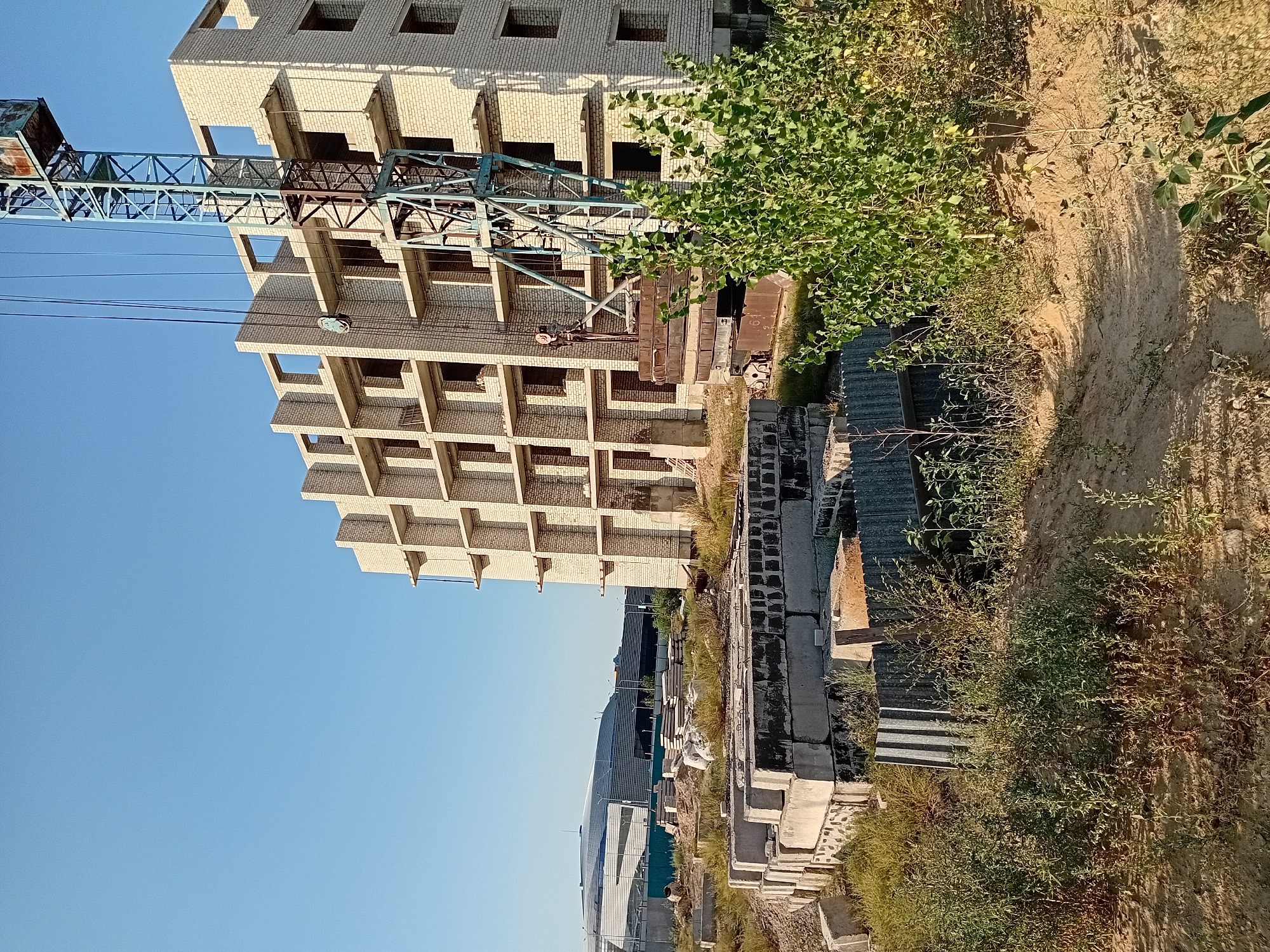 The image depicts an ongoing construction site in the city of Semey, Kazakhstan. A partially constructed multi-story building, made of light-colored brick, dominates the center of the frame. The building has multiple floors with open window and door frames. A large blue construction crane with visible cables is positioned next to the building, suggesting active construction work. In the foreground, stacks of concrete blocks and other construction materials are visible, along with overgrown vegetation and dry earth. In the background, a large modern building with a curved, reflective roof can be seen, possibly a stadium or exhibition hall. The sky is clear and blue, indicating a sunny day. There are no people visible in the image.