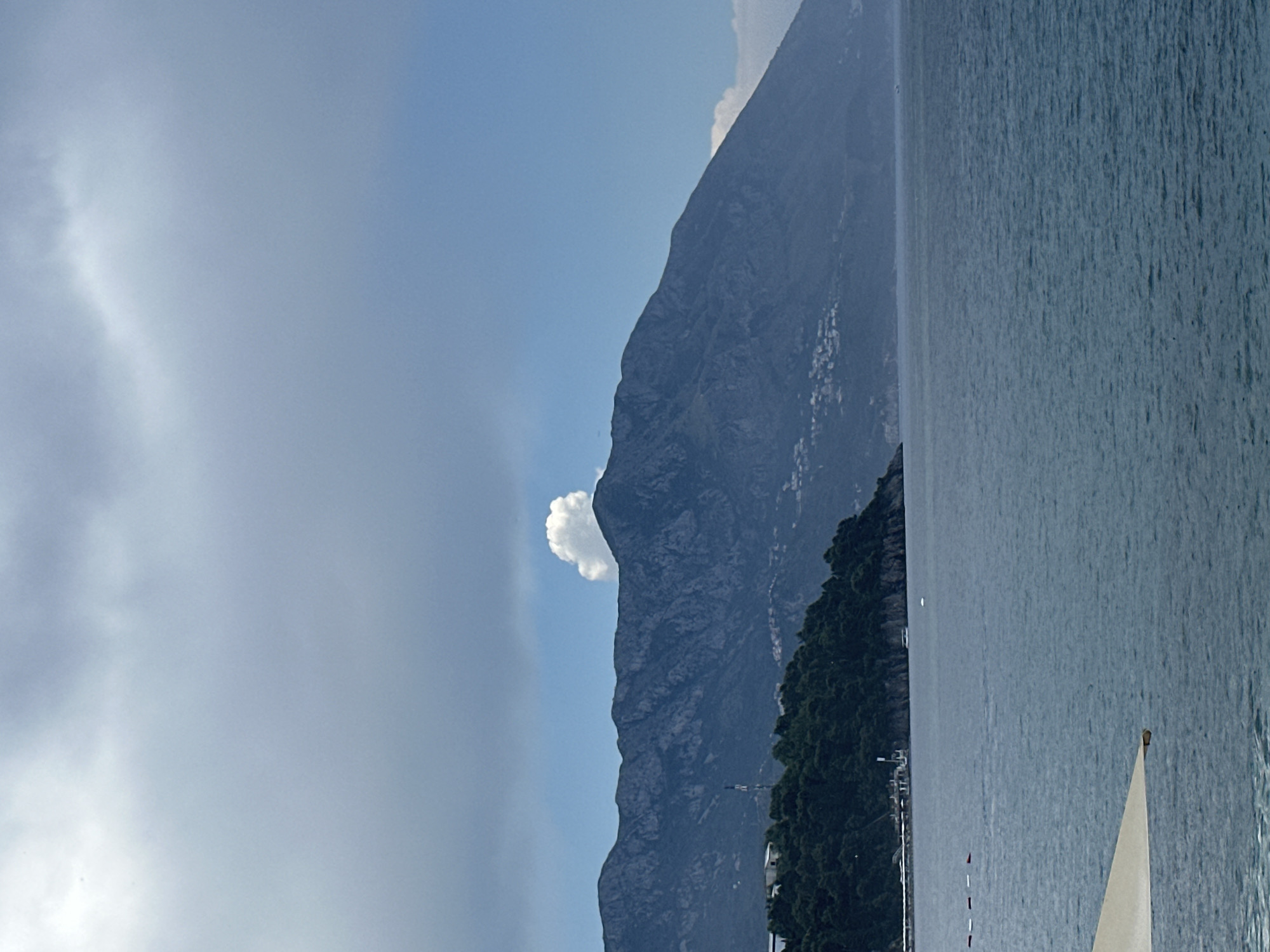 This image captures a panoramic view of the Budva Riviera in Montenegro. In the foreground, the calm, blue sea stretches out towards a distant, tree-covered peninsula. The peninsula is dotted with buildings and features a prominent radio tower. Beyond the peninsula, a vast mountain range dominates the horizon. The mountains are rugged and dark, with a dramatic white cloud formation sitting atop one of the peaks, resembling a mushroom. The sky above is a mixture of blue and grey clouds. A light-colored umbrella is partially visible in the lower-left corner of the frame, suggesting a beach or waterfront setting.