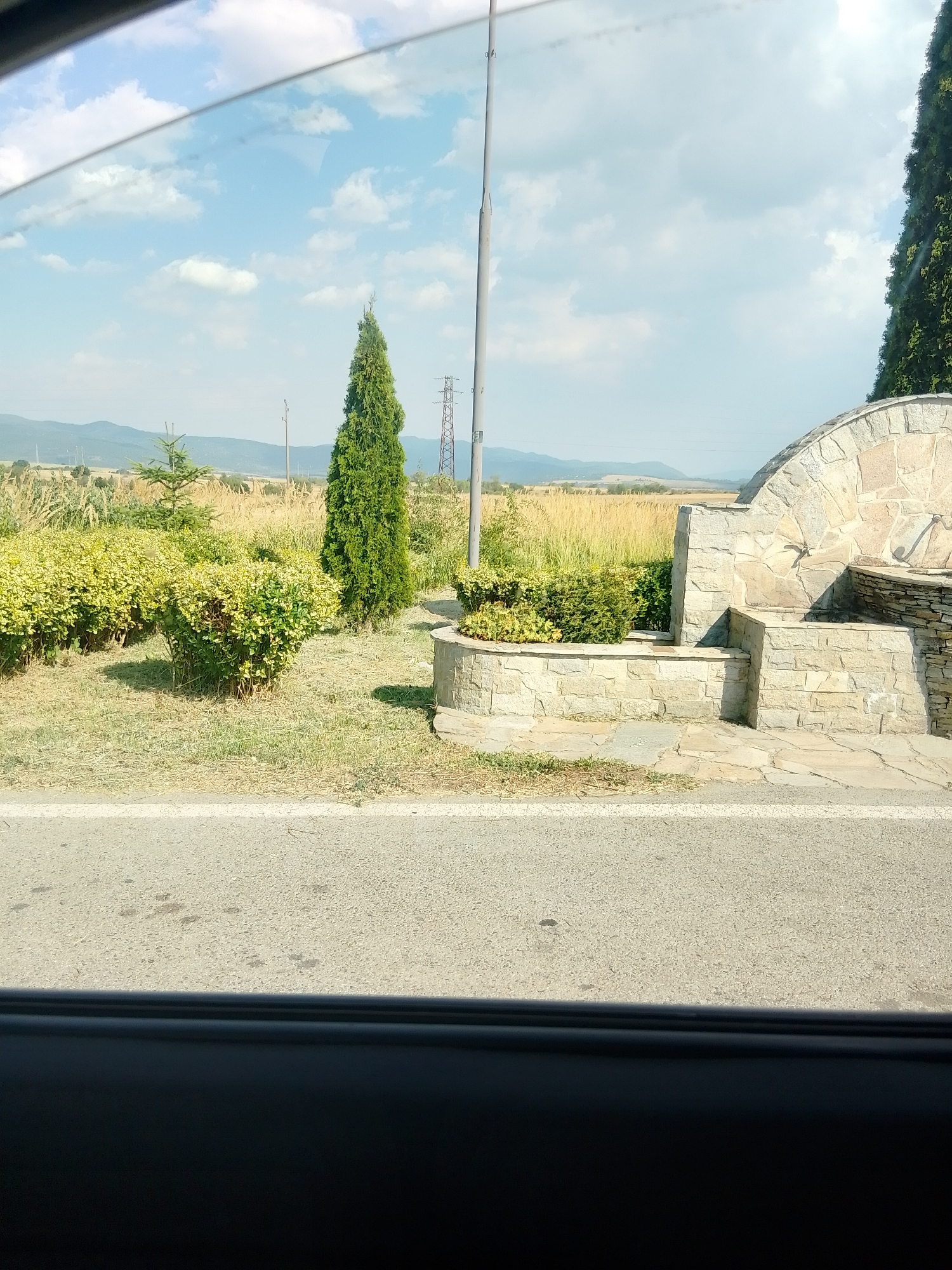The image depicts a roadside view in Kostenets, Bulgaria, on a sunny day with scattered clouds. In the foreground, a paved road with a white line marking is visible, with dry grass and a dark shadow along the bottom edge suggesting the perspective is from within a vehicle. To the right of the road is a low, circular stone structure that appears to be a fountain or decorative element, constructed from rough-hewn stones. Behind this structure, a tall, slender evergreen tree stands out, casting a shadow on the ground.
Beyond the immediate roadside, there is a landscaped area with manicured bushes, some appearing to have yellow-green foliage. Further back, a field of tall, dry grass or wheat stretches towards rolling hills in the distance. A solitary, slender evergreen sapling is planted in front of this field. A tall metal pole, possibly for a light or flag, rises vertically in the mid-ground. In the far background, a power line tower with crisscrossing metal beams is visible against the horizon, along with the faint outlines of distant mountains. The sky is a vibrant blue, marked by fluffy white clouds. The overall scene suggests a rural or semi-rural setting along a road, likely in the outskirts of Kostenets.