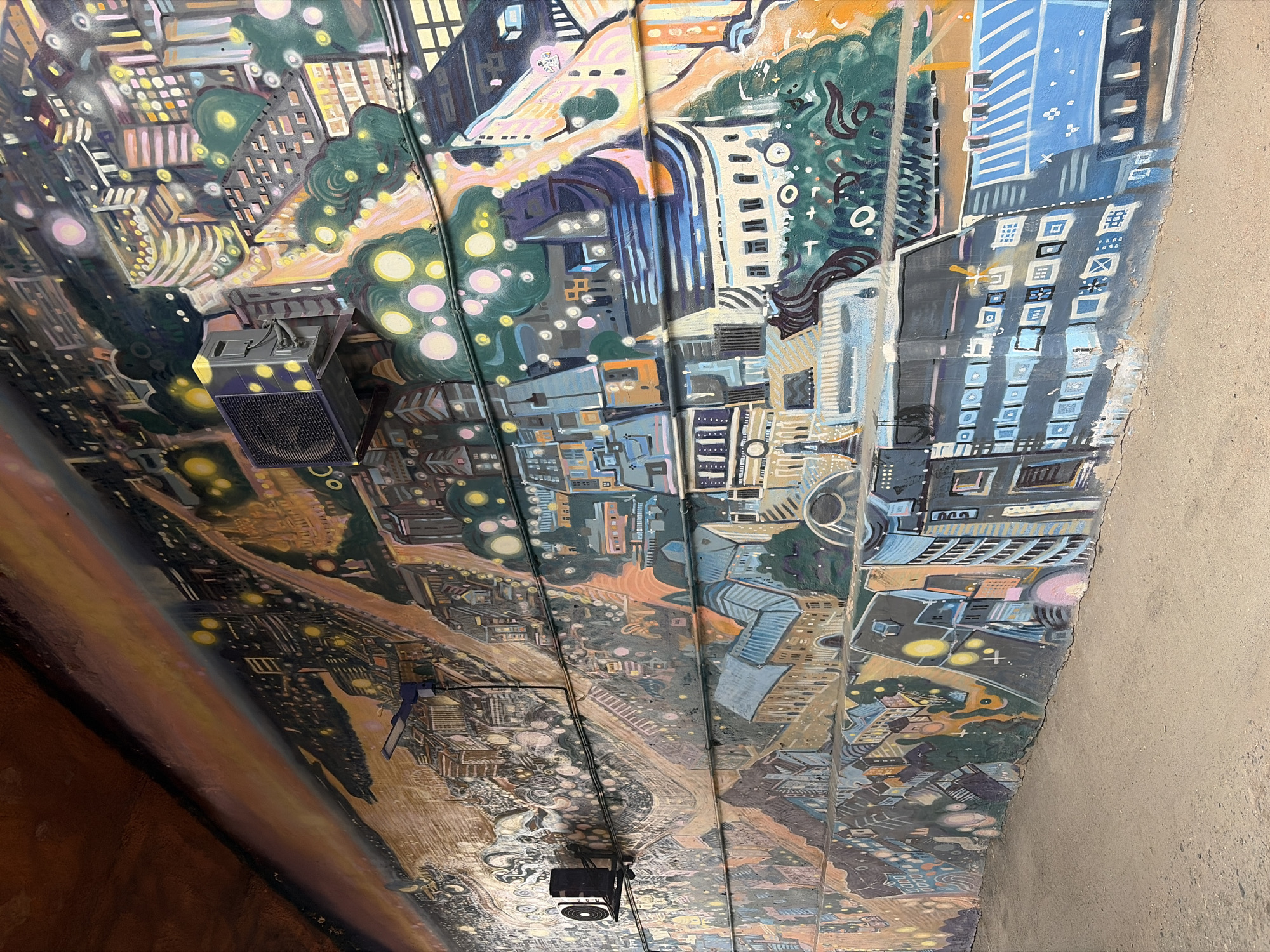 The image shows an urban mural painted on the interior surface of an underpass. The style of the mural is cartoonish with stylized depictions of city buildings, streets, and trees. The color palette is bright with blues, greens, yellows, oranges, and pinks used in the depictions. Several architectural styles of buildings appear to be represented. Overhead infrastructure elements like lighting and wiring are also visible within the painting. The specific location, Izhevsk, Russia, cannot be verified using solely the image, but the architectural style appears consistent with Soviet modernist architecture.