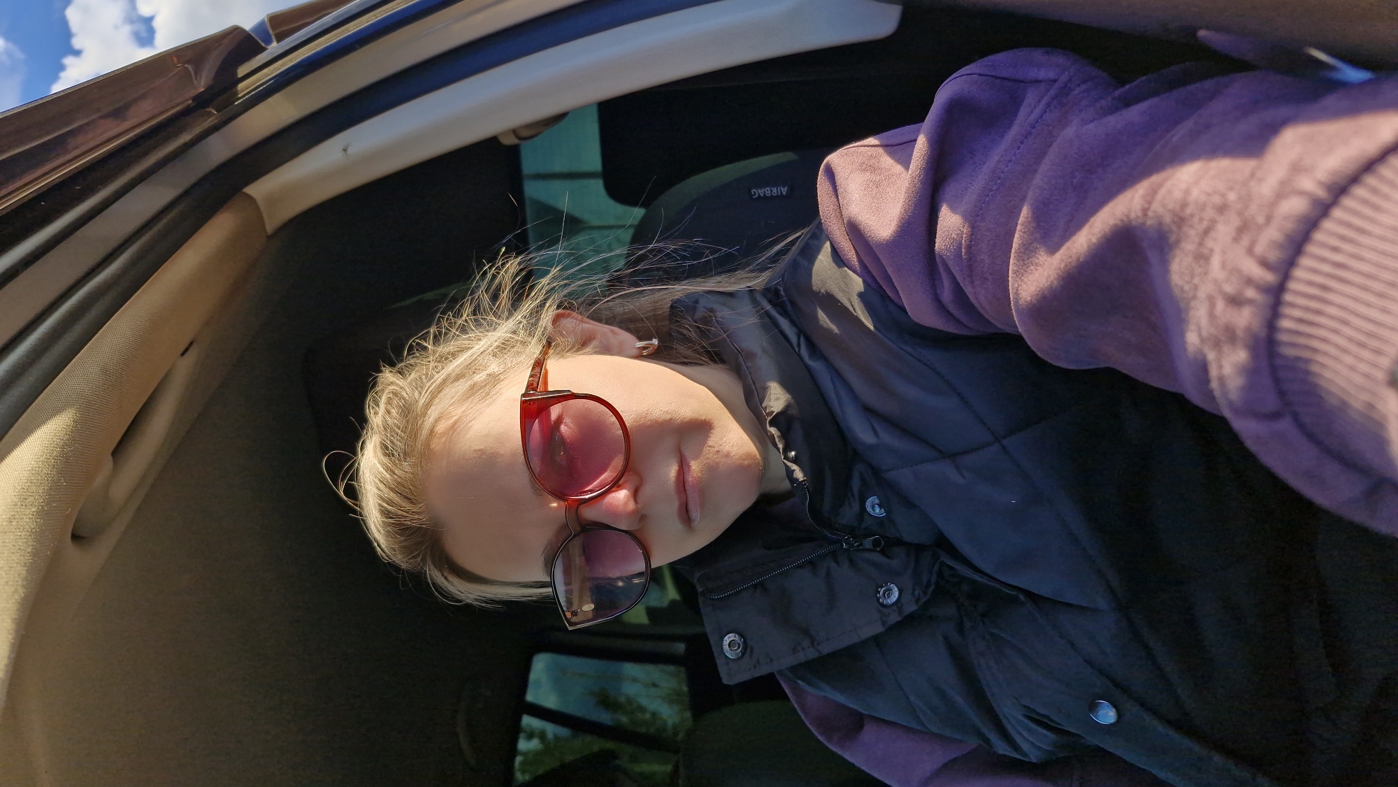 A selfie-style photograph taken inside a vehicle captures a person with light blonde hair and fair skin. The image is rotated counter-clockwise, making the subject appear to be leaning or lying on their side as they look towards the camera from a low angle.
The person is dressed in a purple long-sleeved top, partially covered by a black padded vest. They wear distinctive red-framed sunglasses with reddish-pink tinted lenses that appear to have a darker, lower section, possibly due to reflection or a unique design.
The car's interior is visible, featuring beige fabric on the A-pillar and headliner, and dark upholstery or panels elsewhere. Through a window in the upper left of the frame, a clear blue sky with white clouds is visible, indicating a sunny daytime setting. Strong natural light illuminates the subject's hair and clothing. The word "AIRBAG" is clearly discernible on a dark panel in the background, likely part of the passenger-side interior. The scene takes place in Otchor, Russia.