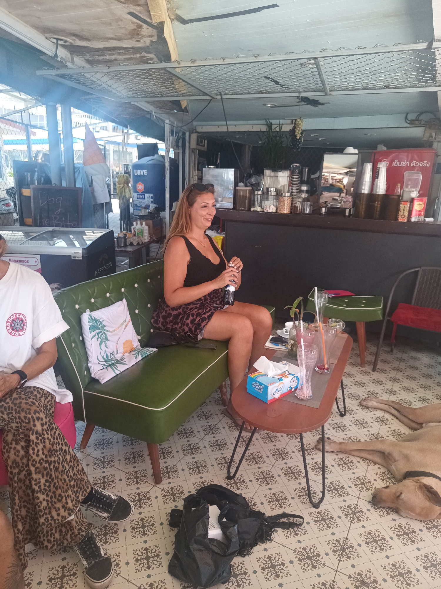 A medium-shot captures the interior of an open-air cafe located in ตำบลเกาะพะงัน, Thailand. A woman with light hair, wearing a black tank top and a leopard print skirt, is seated on a green vintage couch with a white trim, holding a beverage bottle. To her left, another person is partially visible, wearing a white t-shirt with a logo and leopard-print pants, also seated on the same couch. A small round table sits in front of the couch, holding multiple glasses, a small plant, and a blue box of tissues.
On the floor, there's a sleeping dog and a black bag. Behind the woman, a counter stretches across the back of the cafe, holding various jars, bottles, and other cafe paraphernalia. There's also a cooler with signage in Thai. Seating is available along the counter. The floor is covered in patterned tiles, and the lighting suggests a sunny day.