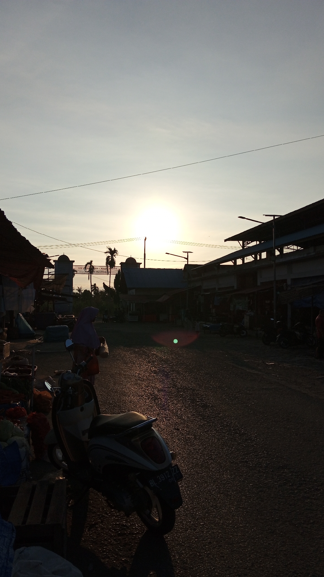 The image depicts a street scene at sunset in Puloara, Indonesia. The sun is low in the sky, casting long shadows and a warm glow over the asphalt road. On the left side of the road, there are market stalls with awnings and various goods, possibly produce, piled on wooden pallets. A scooter is parked in the foreground, its license plate visible.
In the middle ground, a person wearing a headscarf is walking away from the camera, carrying a plastic bag. Beyond them, there are buildings that appear to be part of a market or commercial area, with some palm trees and a decorative archway visible in the distance. The sky is clear with a gradient of pale blue to yellow near the horizon, indicating the setting sun. The overall atmosphere is quiet and tranquil, characteristic of the end of the day in a local Indonesian community.