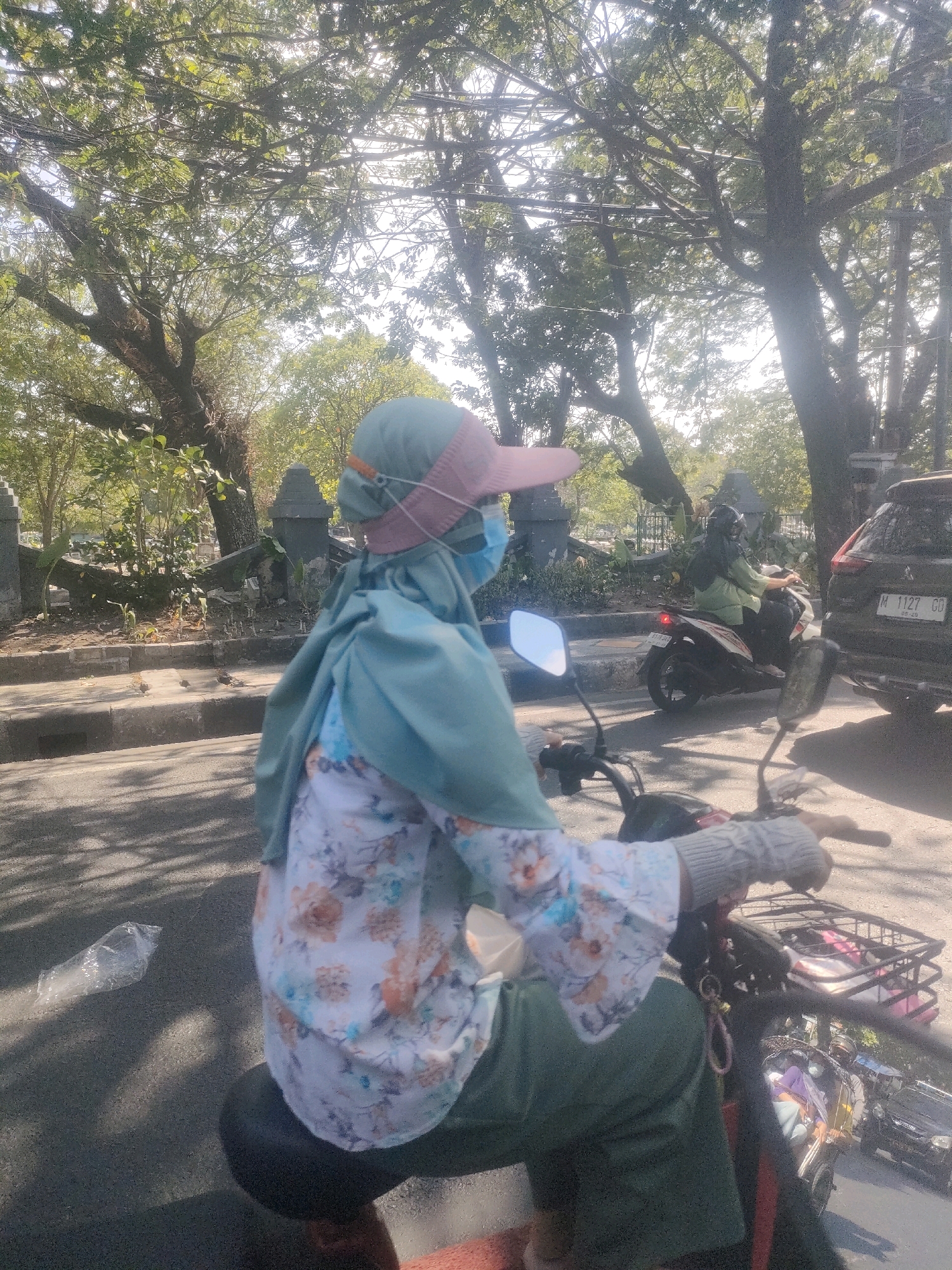 A person wearing a green hijab, a pink visor, and a face mask rides a scooter on a sunny day. The person is wearing a white floral patterned shirt with green sleeves and green pants. The person's right hand is gloved. The scooter has a basket on the front. In the background, there are trees, a cemetery with gravestones, a white motorcycle with a rider, and a black car with the license plate M 1127 CD. The scene appears to be on a street in Surabaya, Indonesia.