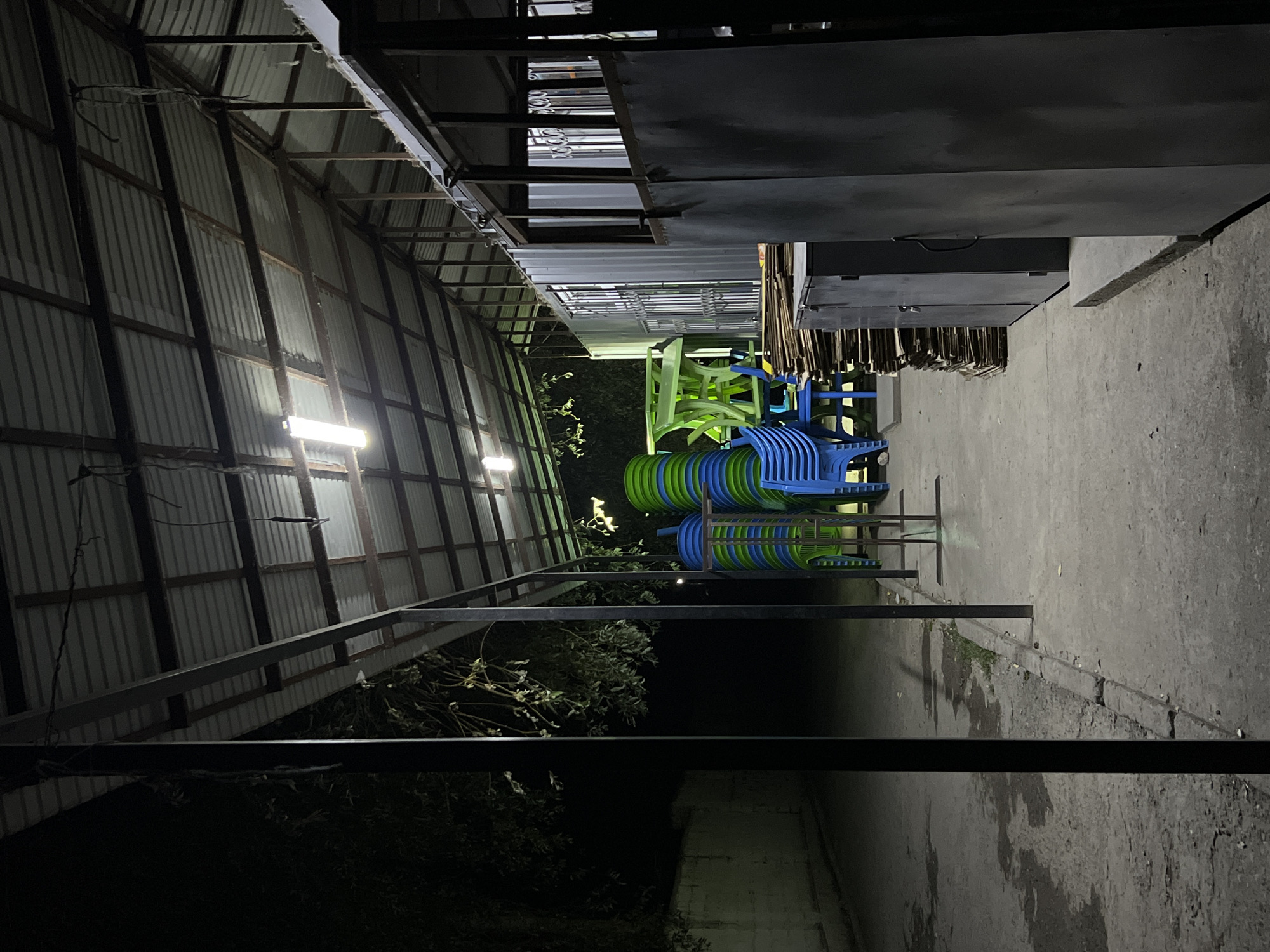 The image depicts a covered outdoor area at night, likely a seating or storage space, in Kadamjay, Kyrgyzstan. The structure has a corrugated metal roof supported by metal beams and poles. Fluorescent lights are mounted on the ceiling, illuminating the space.
On the left side, a paved pathway runs alongside a low white wall. Trees and foliage are visible behind the wall, contributing to a dark, natural backdrop.
In the center of the image, a stack of colorful plastic chairs, predominantly blue and green, are neatly piled. Next to the chairs, a bright green plastic table is also stacked. Further to the right, several tall stacks of cardboard boxes are visible, along with dark gray metal cabinets. These objects suggest storage or preparation for an event.
The overall impression is one of an organized, functional outdoor space used for seating and storage, illuminated for nighttime use. The scene is devoid of any people.