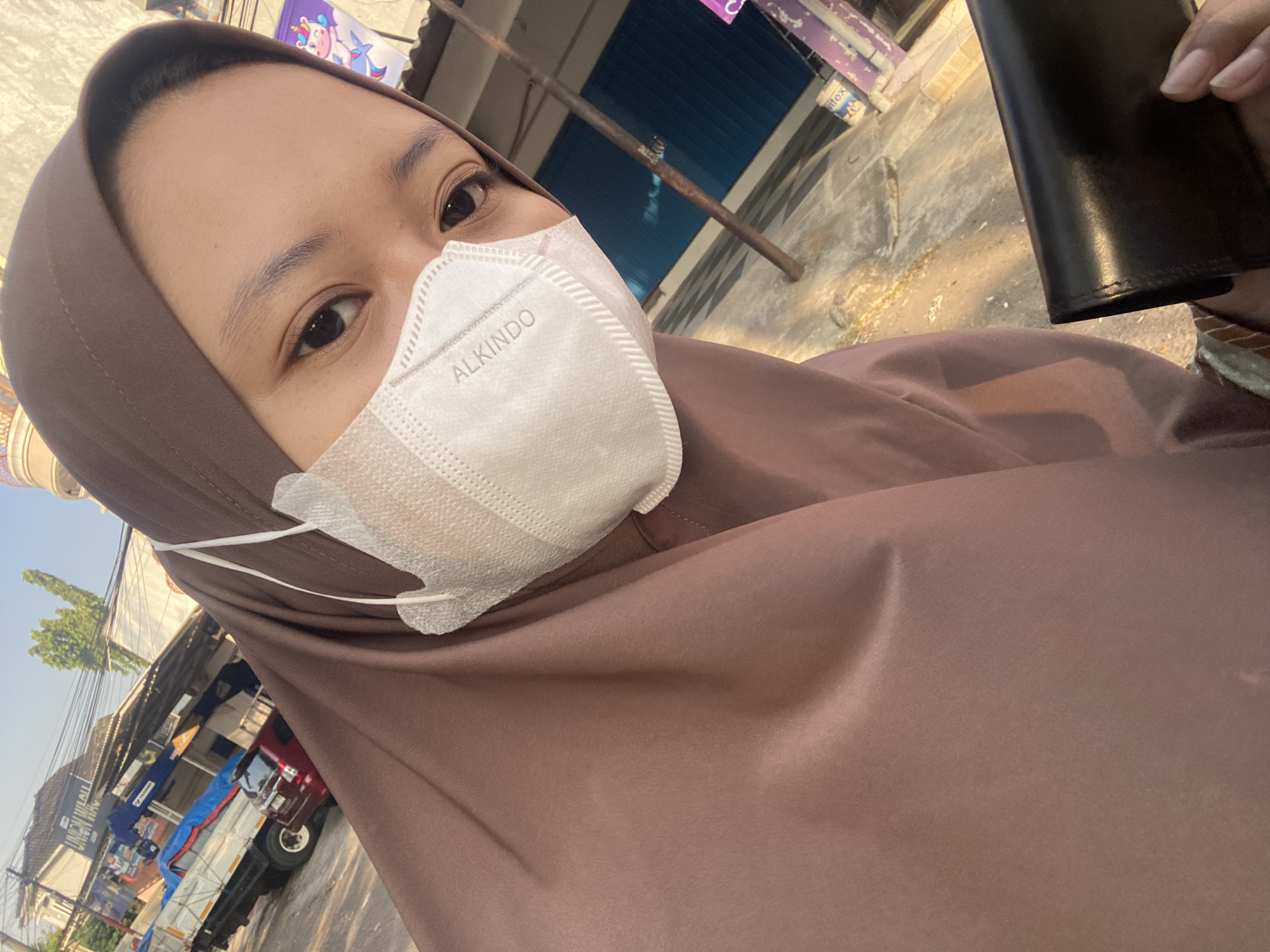 A young woman, wearing a brown hijab and a white face mask, poses for a selfie outdoors on a street. She is holding a black wallet in her right hand. In the background, a red car and a truck with a crane are parked. Buildings with blue shutters line the street. The scene appears to be located in Ngampelsari, Indonesia, given the "UNION WAC" signage visible on one of the buildings and the general aesthetic of the environment. The sunlight suggests it is daytime, likely late afternoon or early morning.
