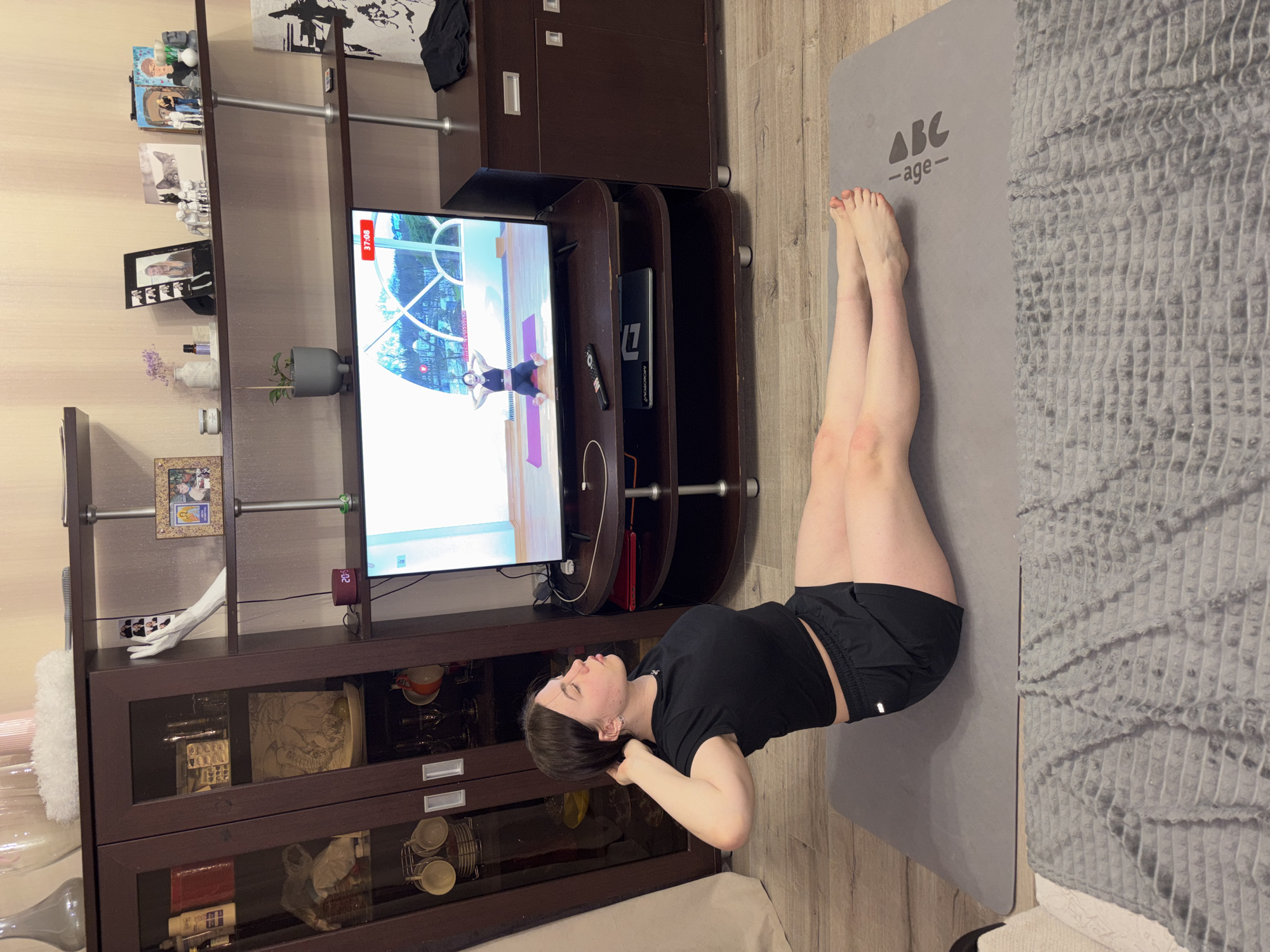 A woman is performing an abdominal exercise on a gray yoga mat in a room with brown furniture. Her legs are raised in the air, and her hands are behind her head. She is watching a video on a television. The room has a cabinet with decorative items and pictures on display. The location is Perm, Russia, based on the timestamp in the television.