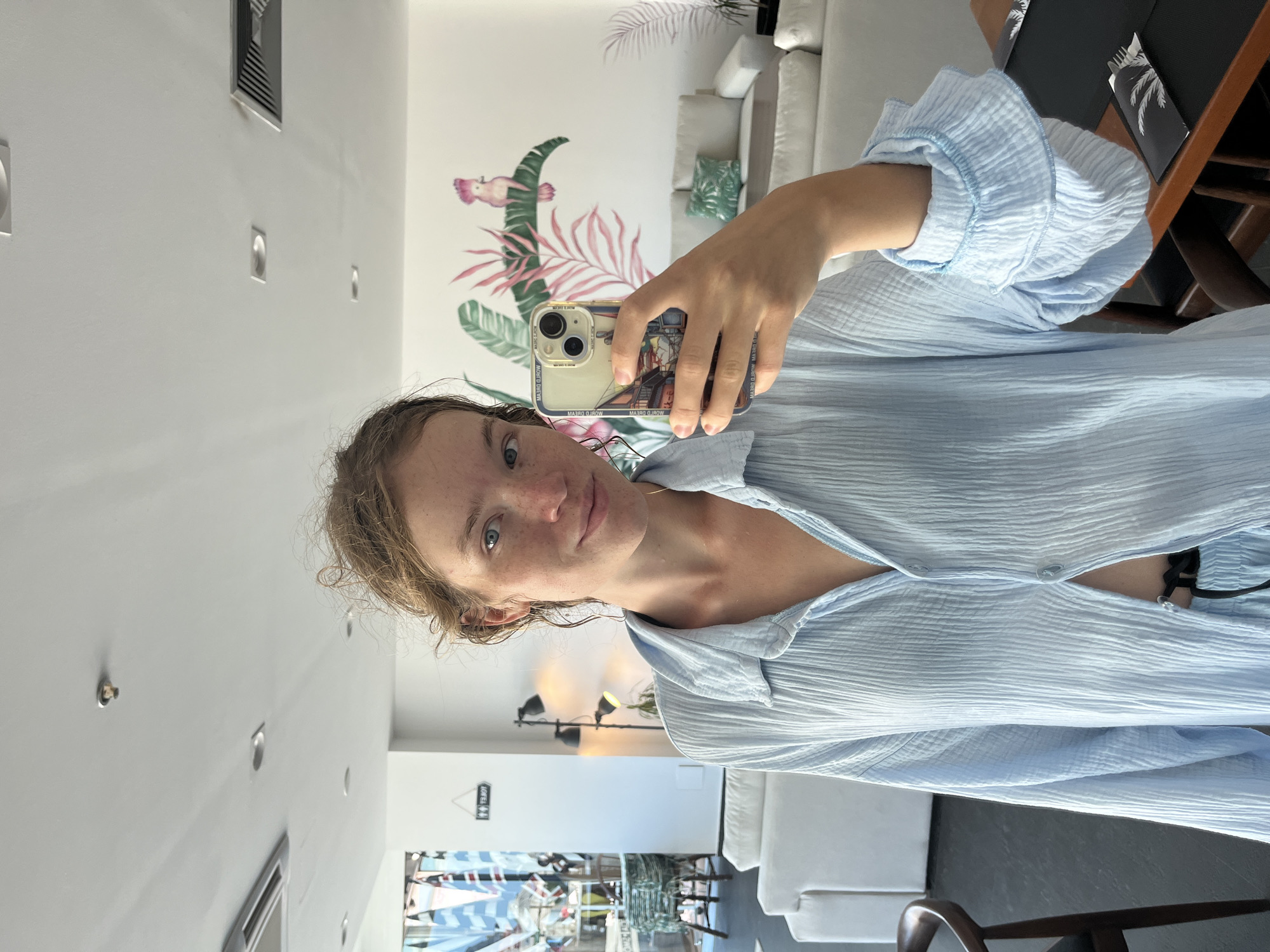 A young woman with freckles and light brown hair, taken from a selfie from the chest up, is wearing a light blue, crinkled button-down shirt. She is looking directly at the camera with a neutral expression. Behind her, a wall is decorated with a tropical mural featuring pink and green leaves and a pink bird. In the background, a white couch and a chair are visible, suggesting an indoor setting such as a living room or lounge. The lighting appears to be natural, possibly indicating daytime. The image is oriented vertically.