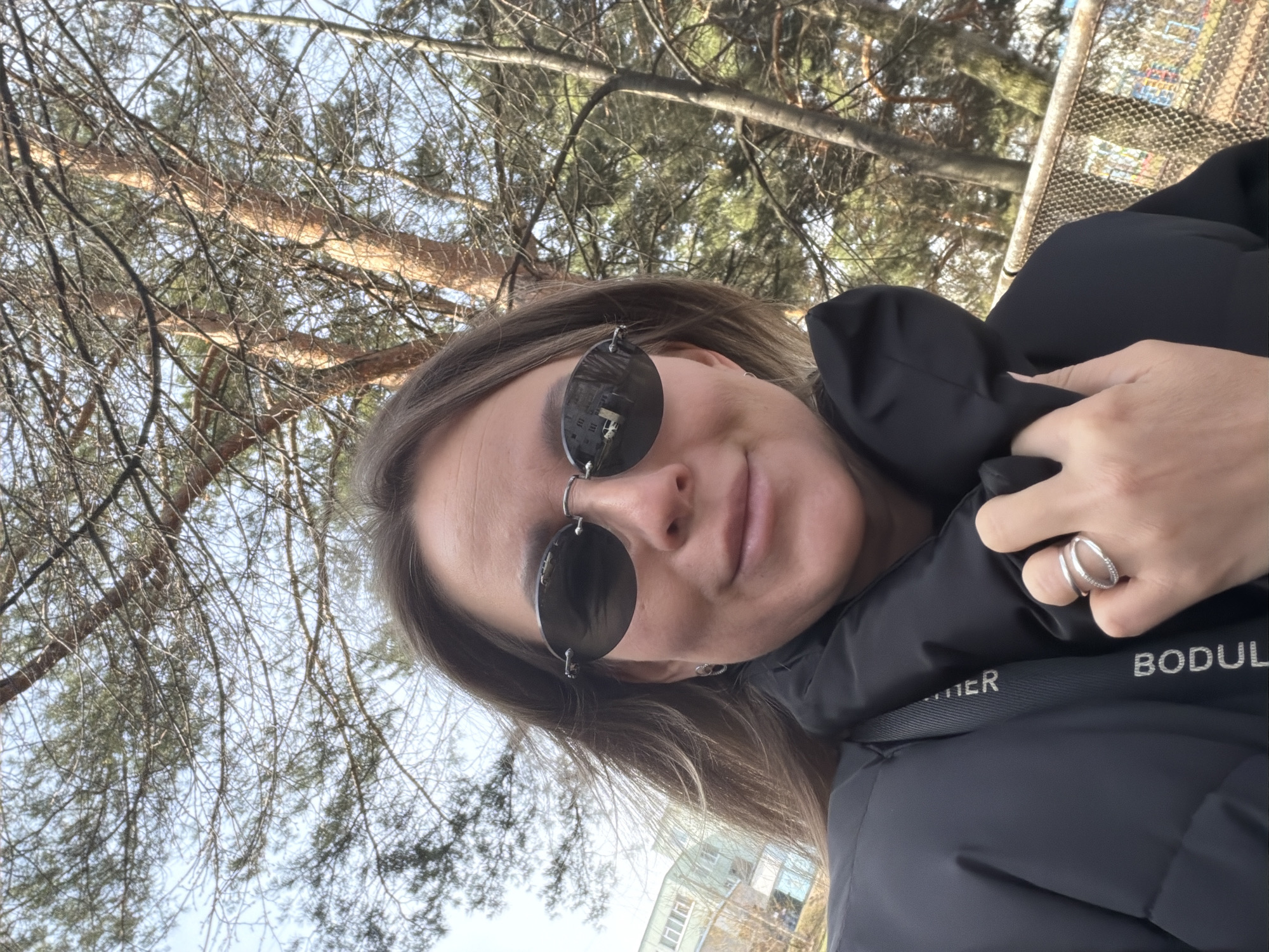 A woman takes a selfie outdoors in Chaikovsky, Russia, captured from a low, upward-looking angle, with the image rotated counter-clockwise. She has light brown hair and is wearing black rimless sunglasses with reflective lenses, which show a reflection of buildings or structures. Her expression is a subtle, closed-mouth smile. She is dressed in a black puffer jacket. Her right hand, adorned with a silver double-banded ring on the ring finger, is visible near her collar, holding her jacket.
The background predominantly features numerous pine trees with long, thin branches, some appearing bare, set against a bright, clear blue sky. The daylight suggests a morning or afternoon setting. A metal fence or railing is partially visible in the upper right background. White text is visible on the black jacket's collar, with "BODUL" clearly legible, followed by a partially obscured "TER" or "TTER." The scene depicts a casual outdoor moment.