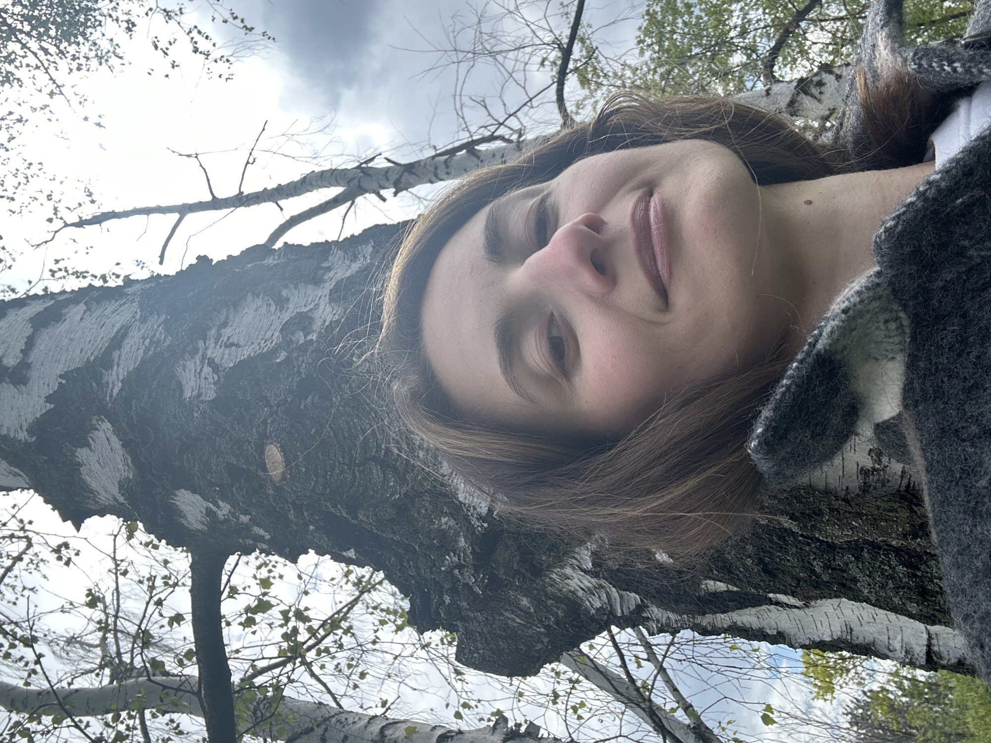 A woman with light skin, brown hair, and wearing a gray and white textured garment lies on a birch tree trunk. The tree trunk shows the characteristic white bark. Above and around the tree, other branches are visible against a cloudy sky. Green leaves appear in the upper right and lower portions of the image. The scene appears to be in an outdoor, possibly forested area. It is not possible to determine the precise location within Kryvyi Rih from the provided image.