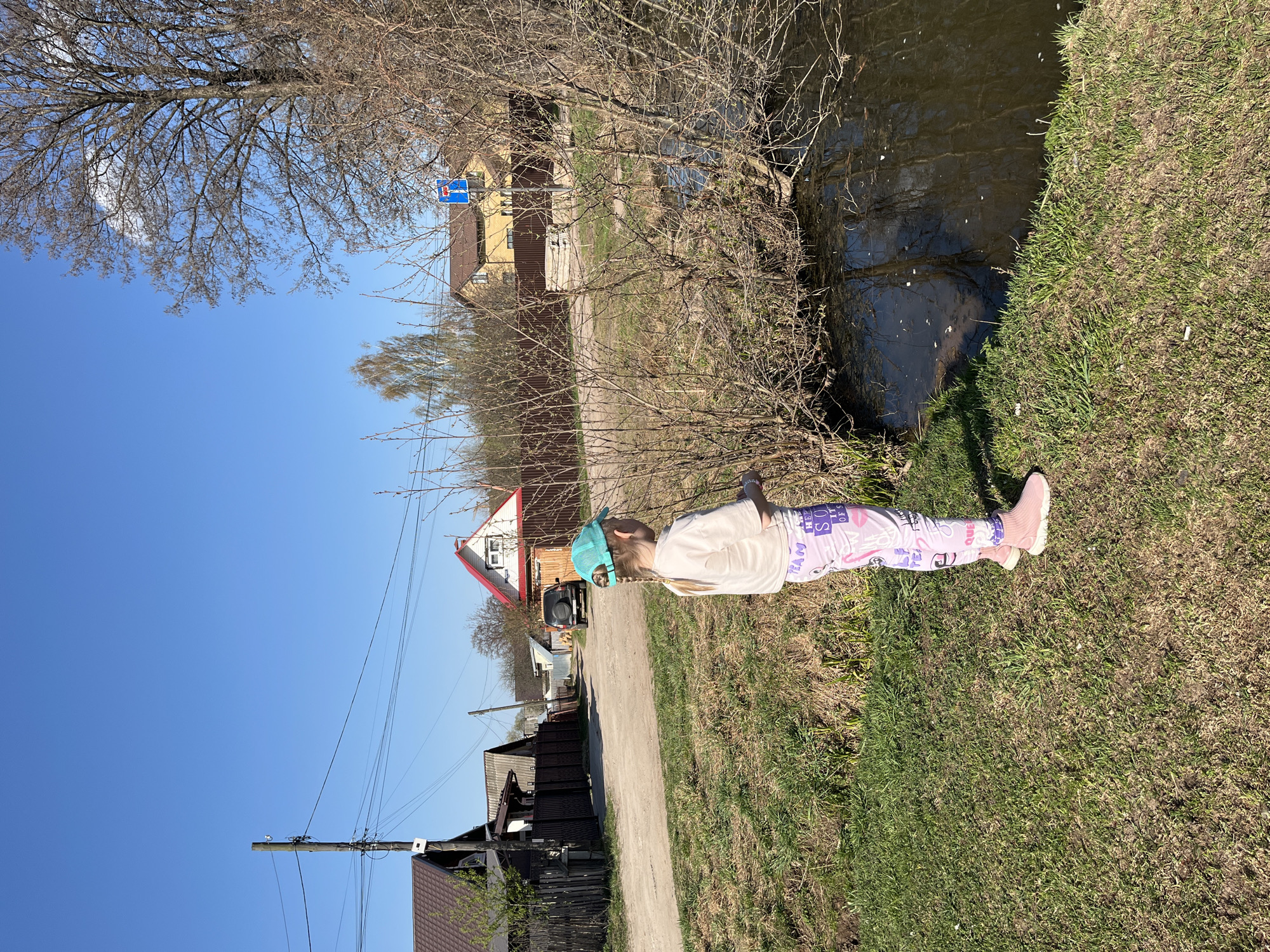 A young girl lies on the grassy bank of a small body of water in what appears to be a rural or suburban area. She is wearing a light-colored sweatshirt, patterned leggings, and pink sneakers. She has a light blue cap on her head. The water is dark and reflects the surrounding vegetation. Behind the girl, a dirt road leads away from the viewer, lined with houses of varying styles and materials. Overhead, power lines run along the road. The sky is clear and blue. The location appears to be in Chaikovsky, Russia, based on architectural styles and environmental characteristics.