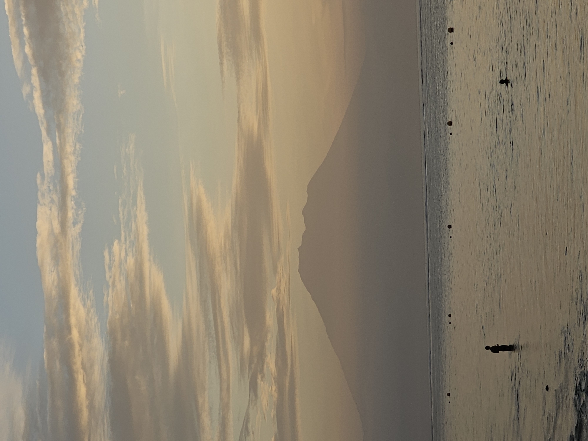 The image depicts a serene seascape with a prominent volcano silhouette in the distance. The sky is filled with soft, pastel-colored clouds, suggesting either sunrise or sunset. The water is calm, with a few buoys scattered across its surface. In the foreground, a lone figure stands in shallow water, silhouetted against the muted light. Another partially submerged figure is visible further out in the water. The overall impression is one of tranquility and natural beauty. Given the presence of a volcano and its proximity to a coastal area, this scene is likely captured on the island of Bali, Indonesia, which is home to several active volcanoes. While Denpasar is the capital city of Bali, it is primarily an urban center and this landscape suggests a more natural coastal setting, possibly on the eastern or northern shores of the island, where volcanic peaks like Mount Agung are visible from the coast.