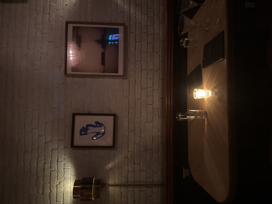 The image displays the interior of a dimly lit establishment, likely a bar or restaurant, located within the city of New York, United States of America. No specific address or landmark for precise location identification is present.
The scene features a light-colored, rounded-edge table in the foreground, set against a white-painted brick wall. A dark banquette or booth seating runs along the wall below a wooden rail. Illumination is provided by a lit candle in a clear glass votive holder centered on the table, casting warm light and reflections. A modern, multi-tiered metallic wall lamp is visible on the left side of the wall.
On the table, from left to right, are two tall clear drinking glasses, one appearing full of liquid. To the right of the candle are two dark rectangular items, likely menus. Further to the right are two empty martini glasses and a smaller clear glass.
Two framed artworks hang on the brick wall. The left frame contains a blue abstract depiction of a reclining or seated figure. The right frame holds a photograph or print exhibiting a soft pink hue, with a prominent blue reflection, possibly from a screen or neon sign, visible in its lower right quadrant. No people are visible in the frame.