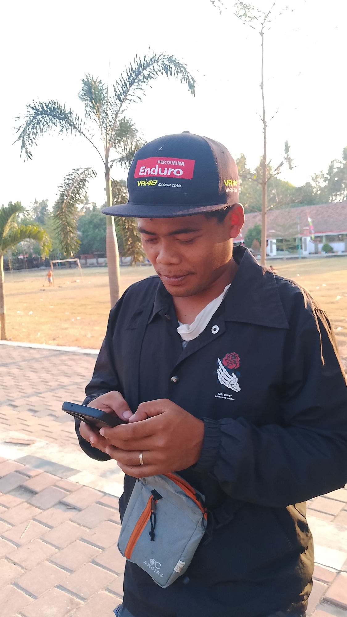 A young man with a dark baseball cap and black jacket is looking down at his smartphone in his hands. The cap has a red and black logo with "PERTAMINA Enduro" and "VR46 RACING TEAM" printed on it. He is wearing a light-colored undershirt beneath his open jacket. A small gray and orange pouch is attached to his belt. He is wearing a ring on his left hand. The background shows a park-like area with trees, a building, and a playground in the distance. The ground is paved with brick-like stones. The lighting suggests it is late afternoon. The image appears to be taken in Karanglangu, Indonesia.