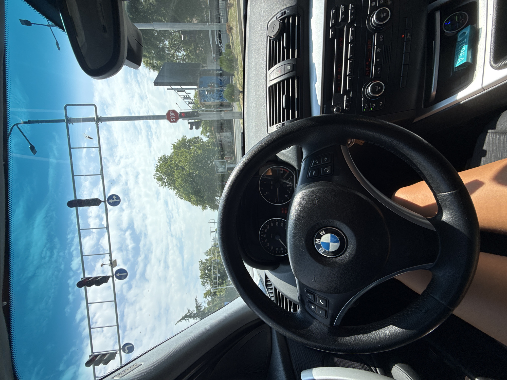 The image shows the interior of a car, likely a BMW given the logo on the steering wheel, viewed from the driver's perspective. The driver's legs are visible below the steering wheel. Outside the car, through the windshield, a sunny sky with scattered clouds is visible. Traffic lights and a stop sign are prominent. The scene appears to be at a traffic intersection. The location is identified as being in the city of Plovdiv, Bulgaria.