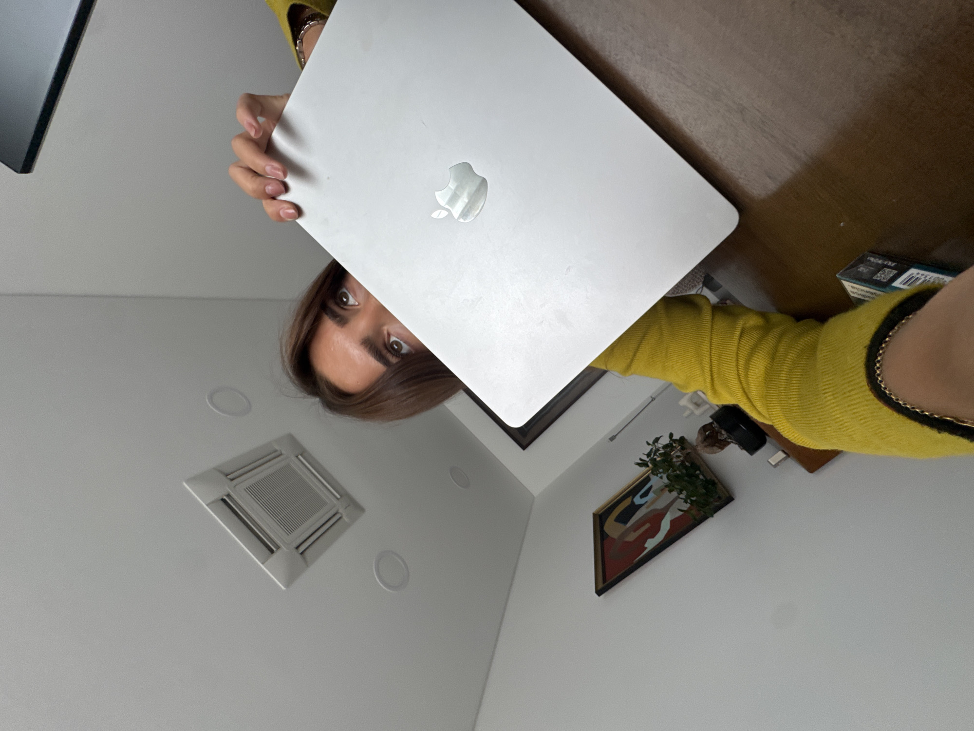A woman holds a closed silver Macbook laptop in front of her face, obscuring most of her features except for her eyes and some of her hair. She is wearing a yellow top. A portion of a table is visible to the upper right. A pack of cigarettes sits on the table next to her right arm. The background shows a white ceiling with a square vent and recessed lighting, and a white wall with an framed artwork. The specific location in the city of Torres Vedras, Portugal cannot be determined from the provided image.
