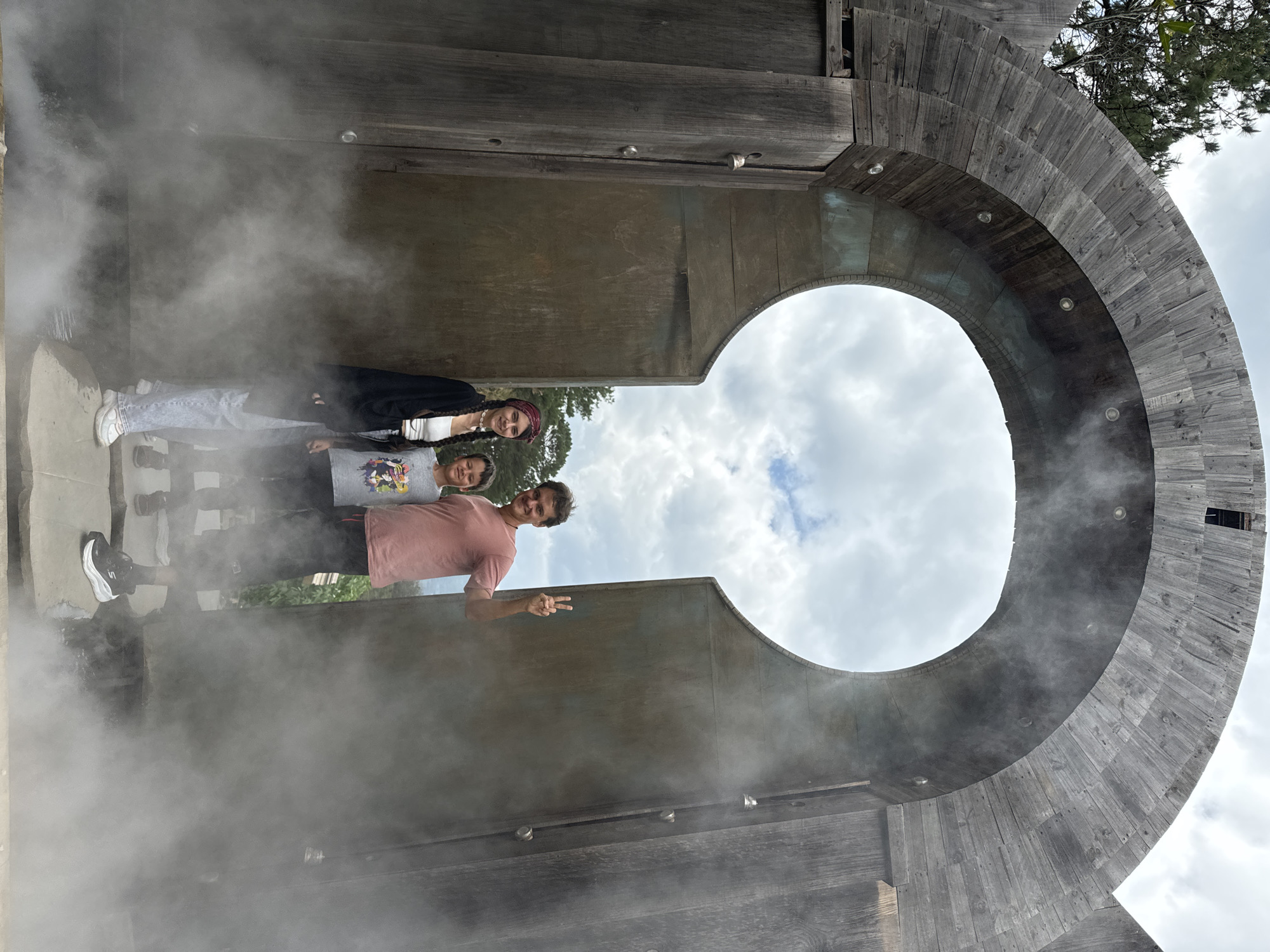 The photo shows three people inside a large, circular wooden structure that resembles a keyhole. The structure is situated outdoors. The atmosphere is filled with mist or steam, obscuring parts of the background.
The individuals are standing close together. The person in the front, an adult male, is wearing a pink shirt and giving a peace sign. Behind him, there is a child wearing a gray shirt. The person furthest back appears to be a young woman with braided hair.
The sky is visible through the keyhole opening, suggesting an overcast day. The wooden structure is made of layered planks and is well-lit. It appears the location is outdoors in the city of Thôn 1, Vietnam.