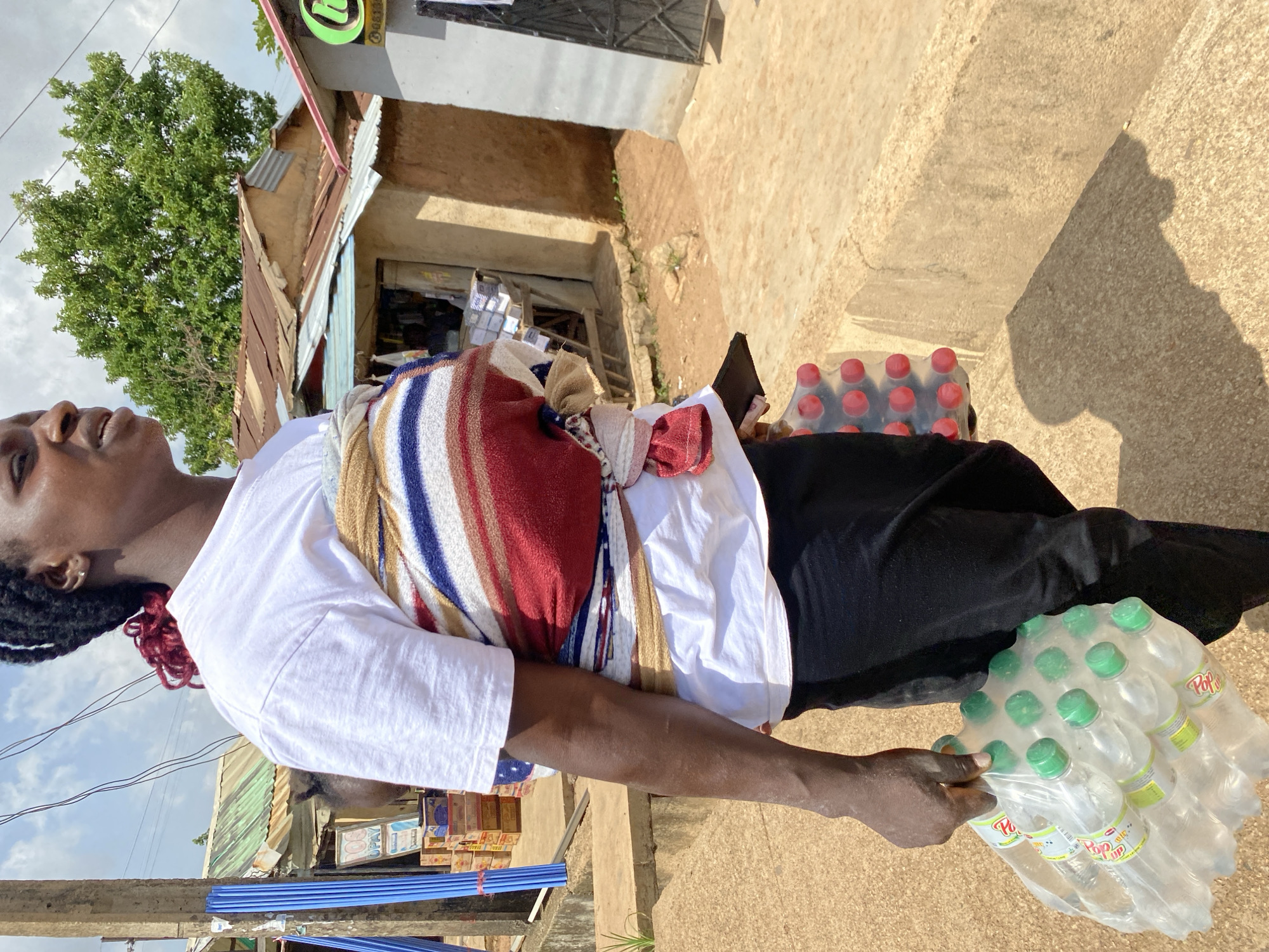 A woman in Bukuru, Nigeria, is captured in a medium shot, from the knees up, carrying a baby strapped to her chest with a colorful cloth. She wears a white t-shirt and black pants. In her left hand, she holds a case of bottled water with green caps, stacked in two rows of five bottles each. The background suggests a market or roadside environment with various buildings, including structures with corrugated iron roofs. A prominent wooden post with blue pipes is visible on the left side of the frame. The ground is a mix of concrete and dirt. The lighting indicates it is daytime, with shadows cast on the ground.