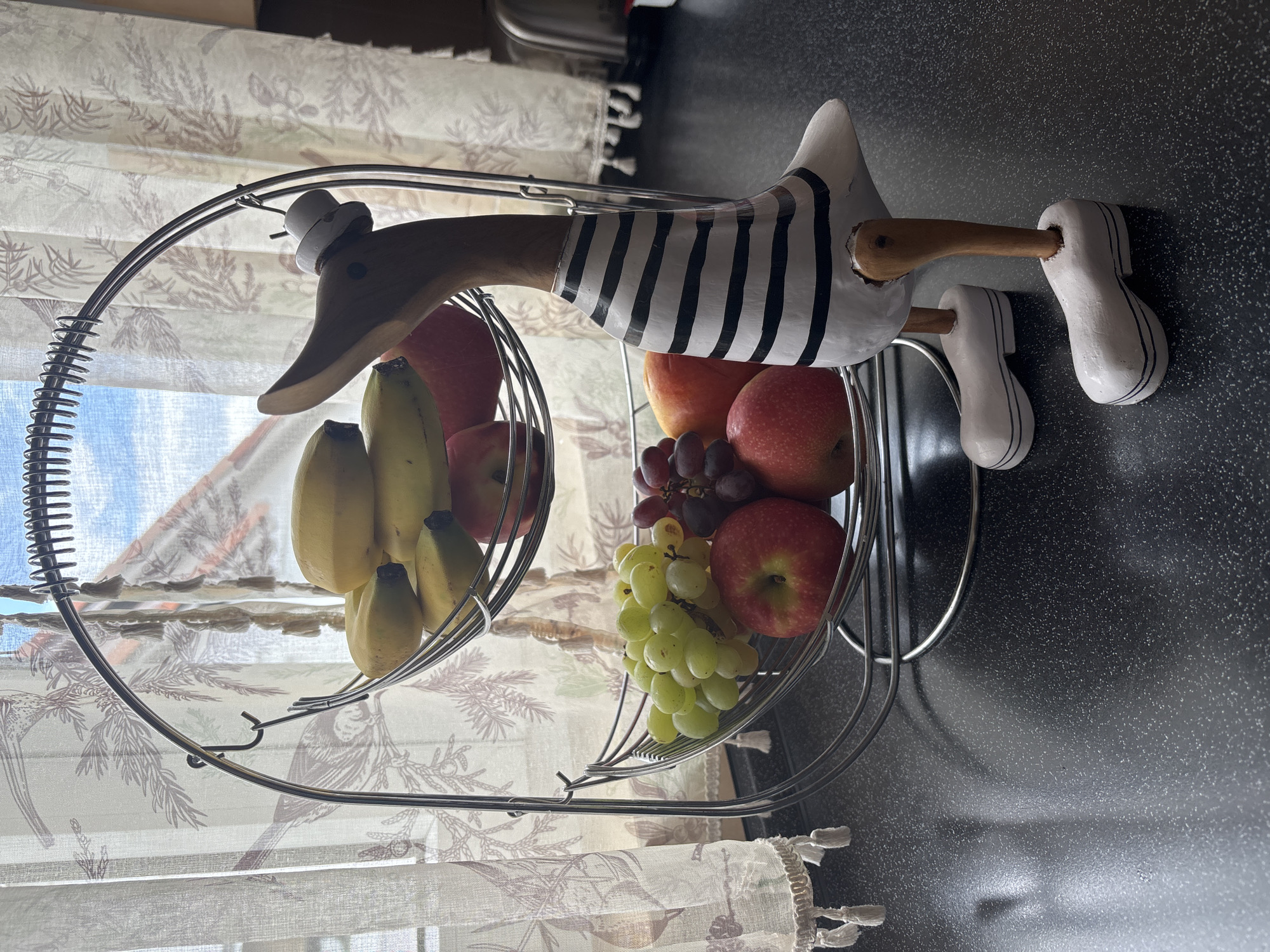 The image captures a two-tiered fruit basket featuring a decorative wooden duck figure, set on a dark, speckled countertop. The scene appears to be an interior space, likely a kitchen or dining area, with natural light entering from a window in the background.
The central subject is a whimsical wooden duck figure, which serves as a handle or decorative element for the fruit basket. Its head and neck are natural light brown wood, while its body is painted white with distinctive black horizontal stripes, resembling a classic sailor's shirt. The duck wears a small, white top hat on its head and has wooden dowel legs ending in white wooden clogs with black striped details. It appears to be holding onto the top bar of the metal fruit stand.
The fruit basket itself is made of silver-colored metal wire. The top tier contains a bunch of yellow bananas and some red apples. The larger bottom tier holds a variety of fruits, including red apples, clusters of green grapes, and dark red grapes, along with what appear to be peaches or nectarines.
In the background, a window is visible, covered by light-colored fabric curtains or roman shades. The curtains feature a repeating pattern of foliage, birds, and possibly keys, and have a decorative fringe or tassel along the bottom edge. Bright diffused light suggests it is daytime. The countertop beneath the fruit basket is dark gray or black with small white speckles, characteristic of a granite or quartz surface. No visible text is present in the image.