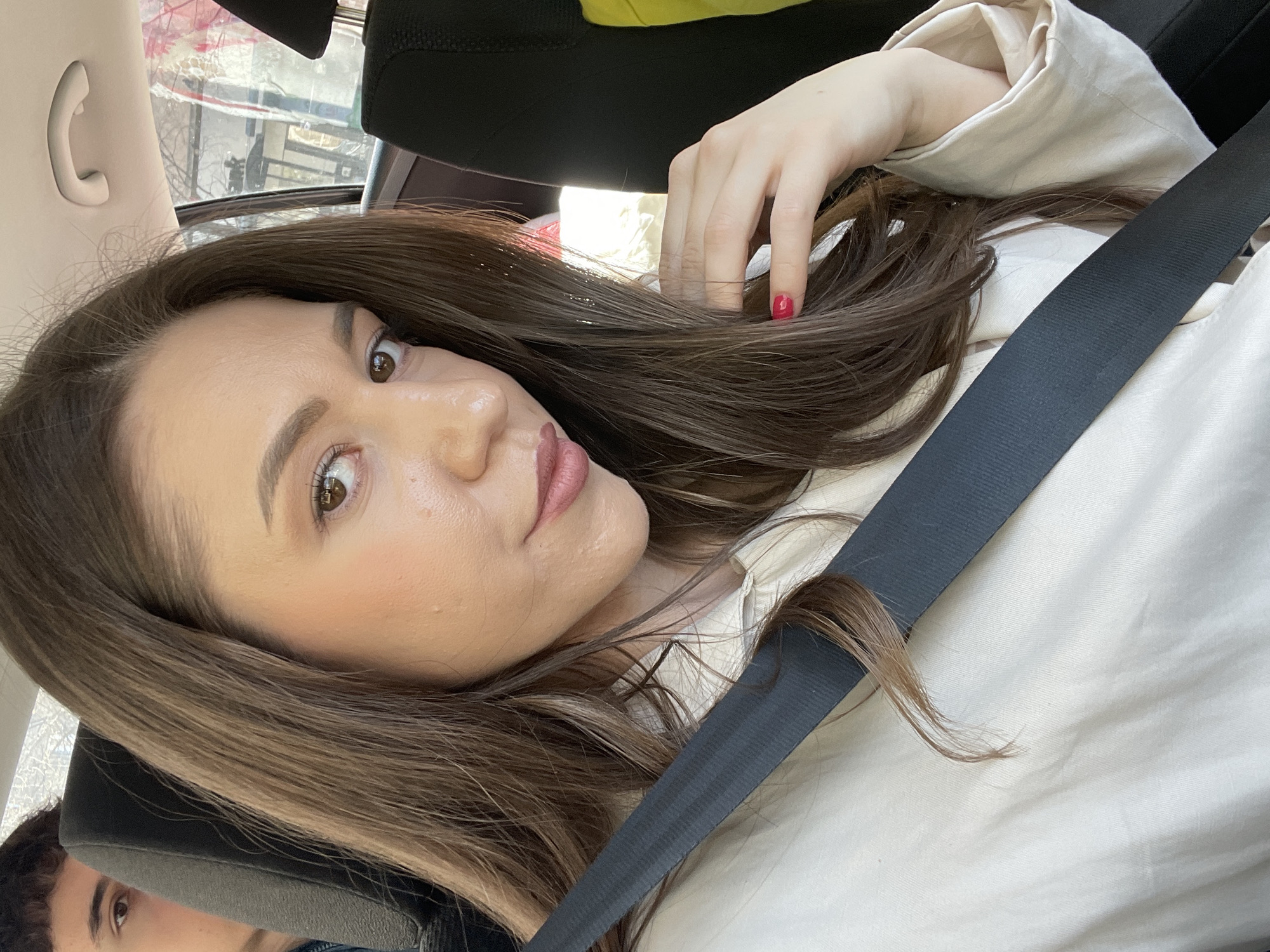 A woman with shoulder-length brown hair is in a car, wearing a seatbelt and a beige shirt. She has a red nail on one finger of the hand near her hair. The photo is taken from an angle and the location might be the city of Varna in Bulgaria, but that's not certain. The car's interior includes a headrest and a window with a view of the outside. A face is visible in the bottom left corner of the image.