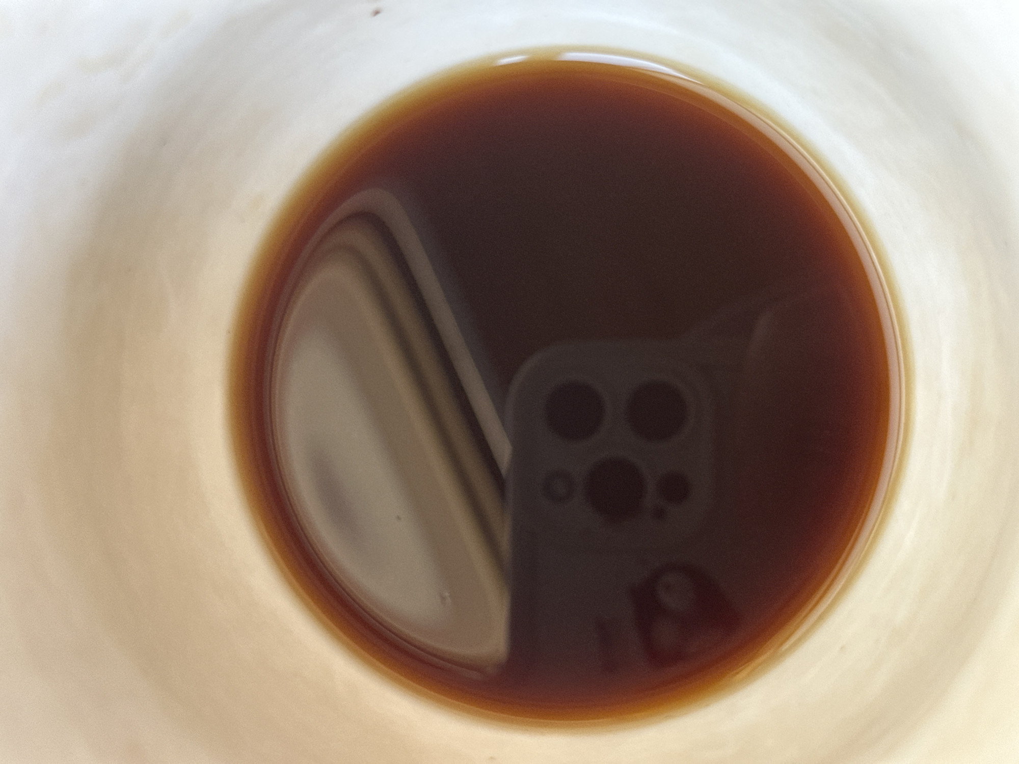 The image shows a top-down view of a liquid, likely coffee or tea, in a light-colored cup. A reflection in the liquid suggests a mobile phone is being held above the cup to take the photo. The phone appears to have multiple camera lenses. The location cannot be determined from the image, and there is no indication of Nizhny Novgorod, Russia.