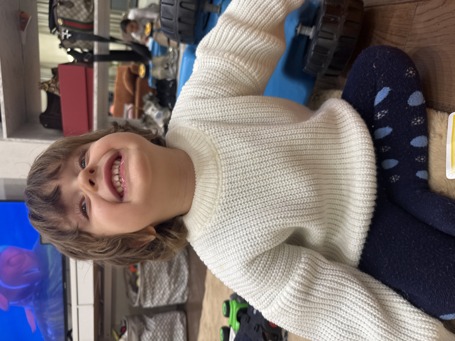A young child in a white sweater smiles broadly. They wear blue tights with light blue polka dots. A blue toy truck is nearby. The background is a room with shelves holding bags and boxes, a television screen showing a fish, and decorative storage containers. A person sits in the background.