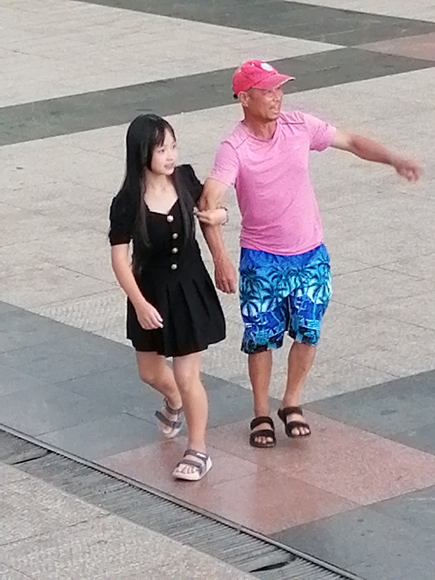 The image shows two people walking outdoors on a paved surface, possibly a plaza or walkway. The person on the left is a young female wearing a black dress and white sandals. The person on the right is an adult male wearing a pink t-shirt, blue patterned shorts, a red baseball cap, and dark sandals. They are walking side-by-side, with the man's arm around the woman's shoulder or upper arm. The ground is made of large, square tiles in varying shades of gray and some pinkish-red. There is a metal grate running along the bottom edge of the image. The lighting suggests it is daytime, with no indication of specific weather conditions. The scene appears to be in a public outdoor space.