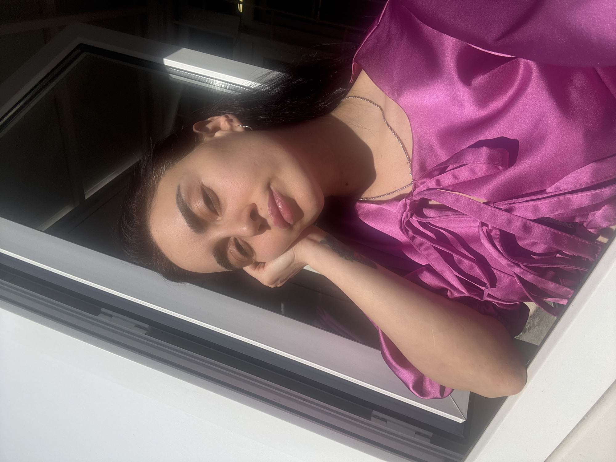 A young woman with dark hair is resting her head on her hand, framed by a white window. She is wearing a bright pink, satiny top with fringe detail. She appears to be looking out the window. The location is not discernible from this image alone and cannot be confirmed as Tbilisi, Georgia.