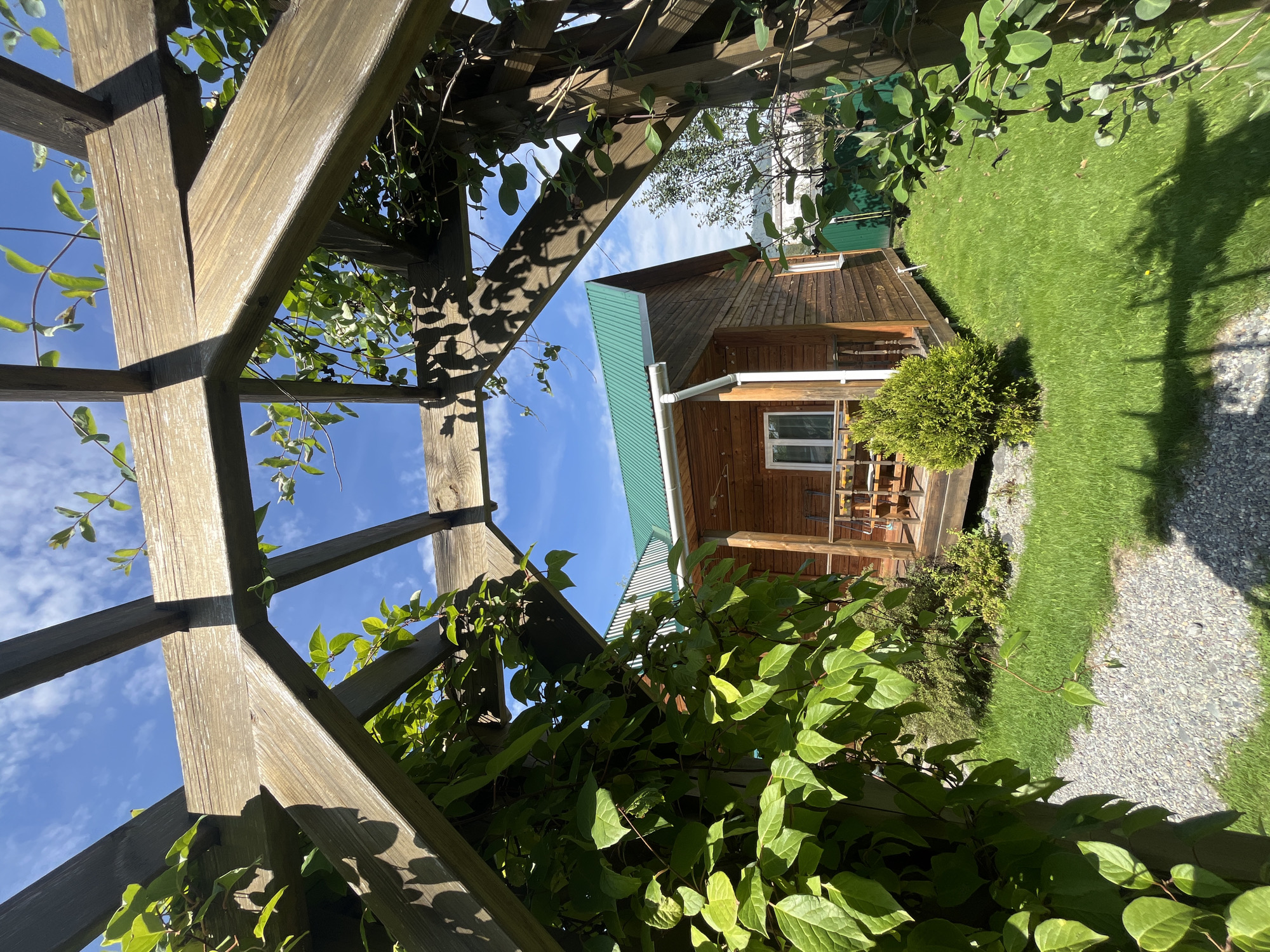 The image shows a wooden house with a green metal roof, situated in a lush green garden. The house has a wooden porch with a table and chairs. The garden is well-maintained, with manicured grass and a gravel path. In the foreground, a wooden pergola with climbing plants partially obscures the view of the house and garden. The sky is blue with scattered white clouds. The scene is set in Ust'-Sema, Russia, during daylight. There are no people visible in the image. The environment appears to be peaceful and idyllic.